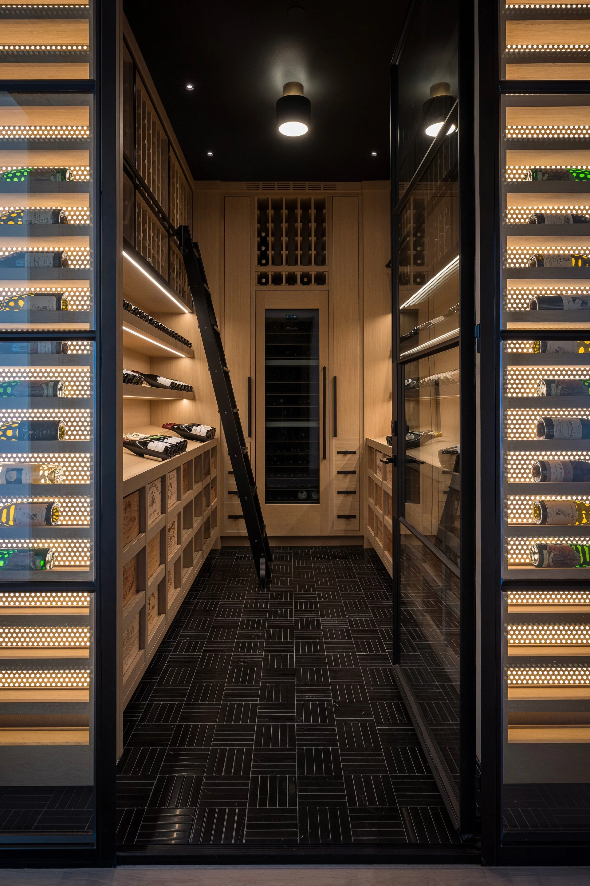 A custom light wood wine cellar features backlit shelves with wine bottles, a black rolling ladder, and dark tile flooring.