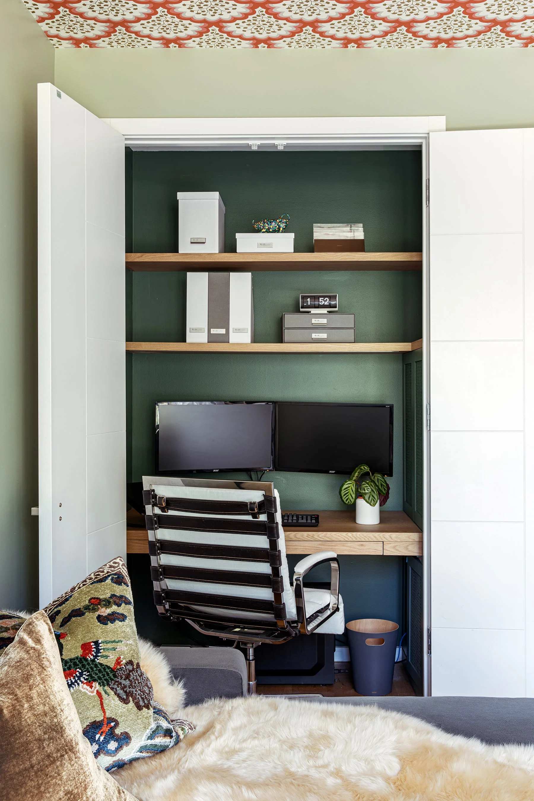 A closet transformed into a compact home office with a built-in wooden desk, floating shelves, and deep green walls.