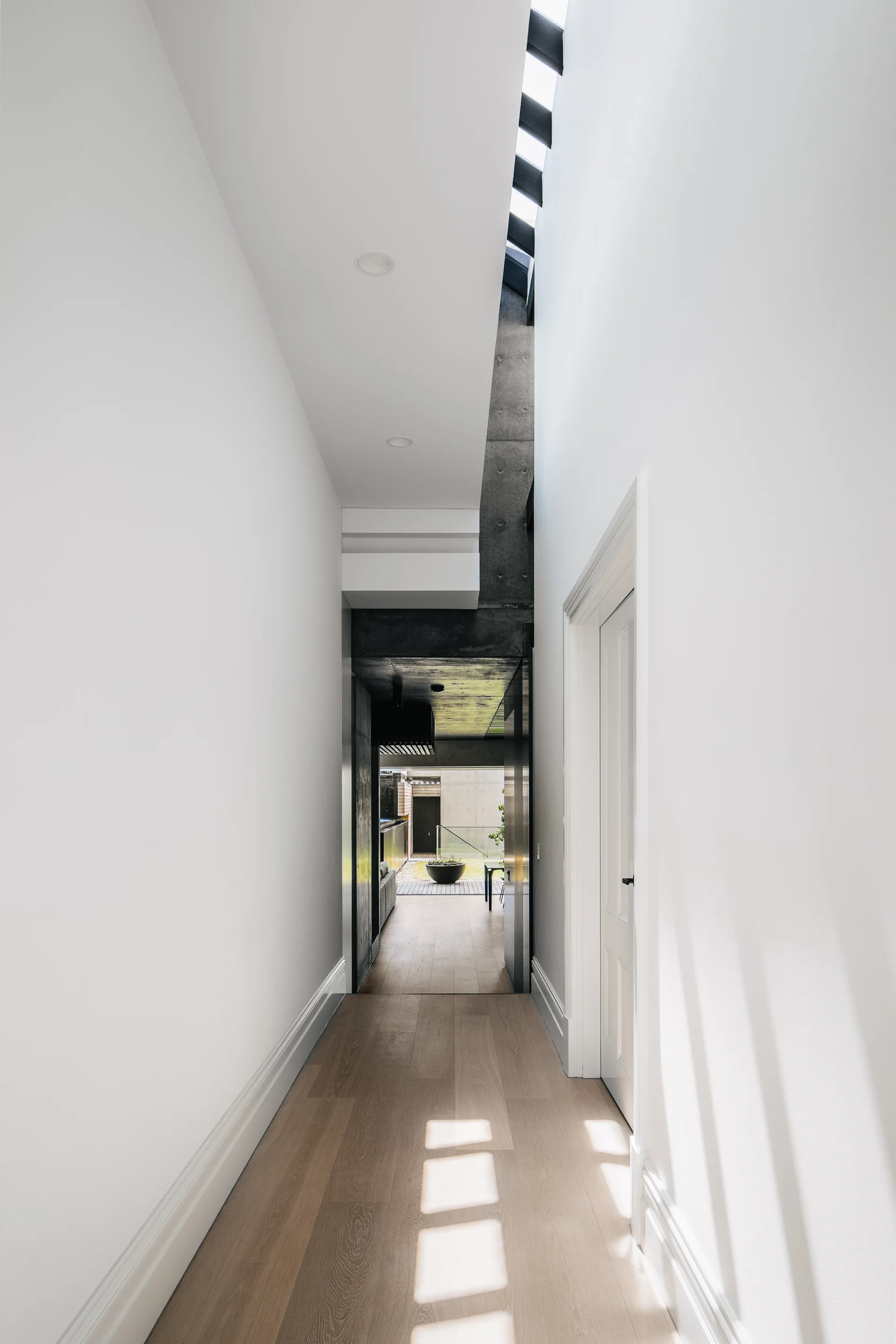 A narrow white hallway with light wood flooring features a long slit skylight on the ceiling and a view toward an outdoor courtyard.