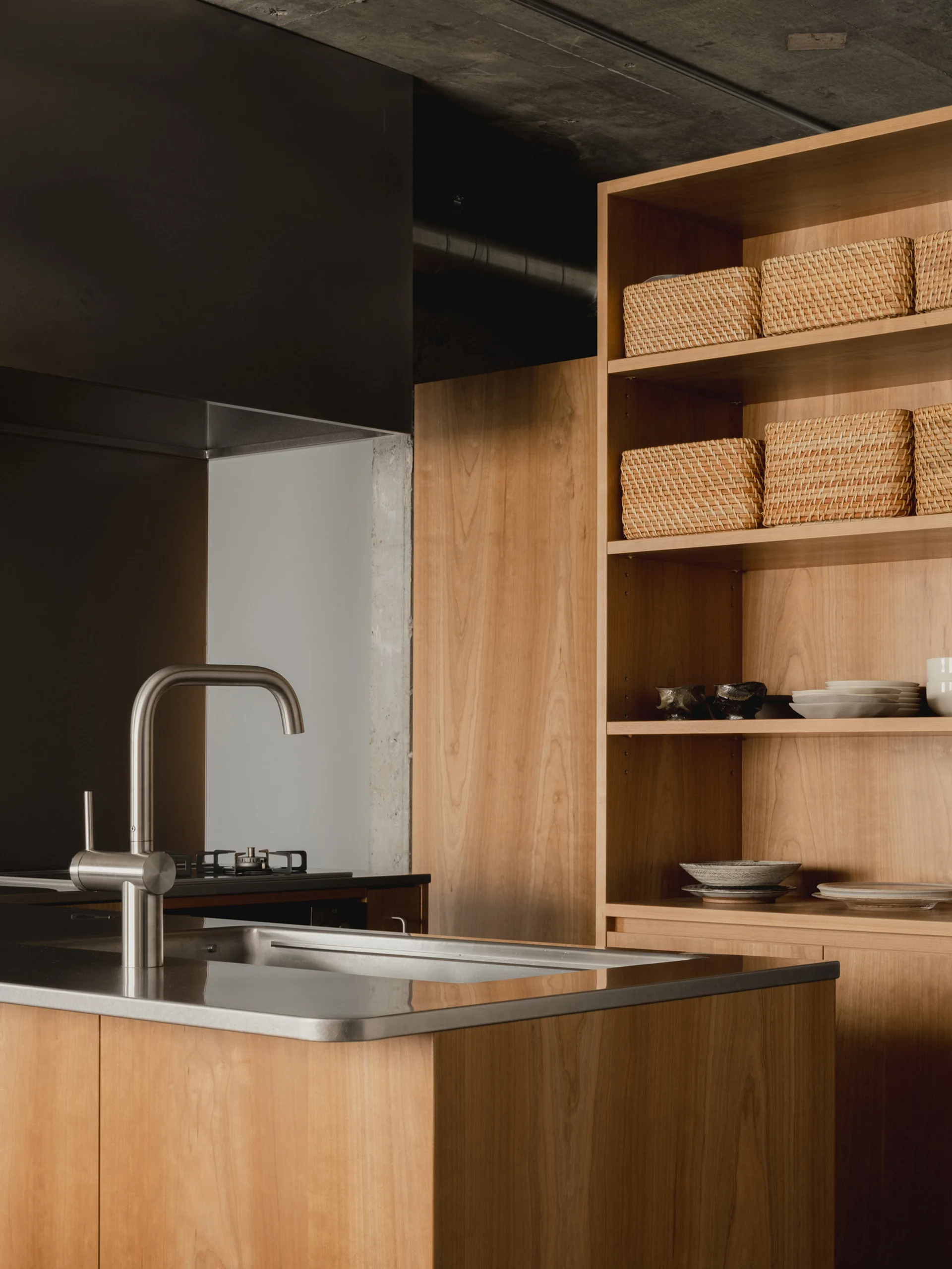 A modern kitchen featuring a wood island with a stainless steel sink, next to tall wooden shelves filled with woven baskets and ceramic dishes.