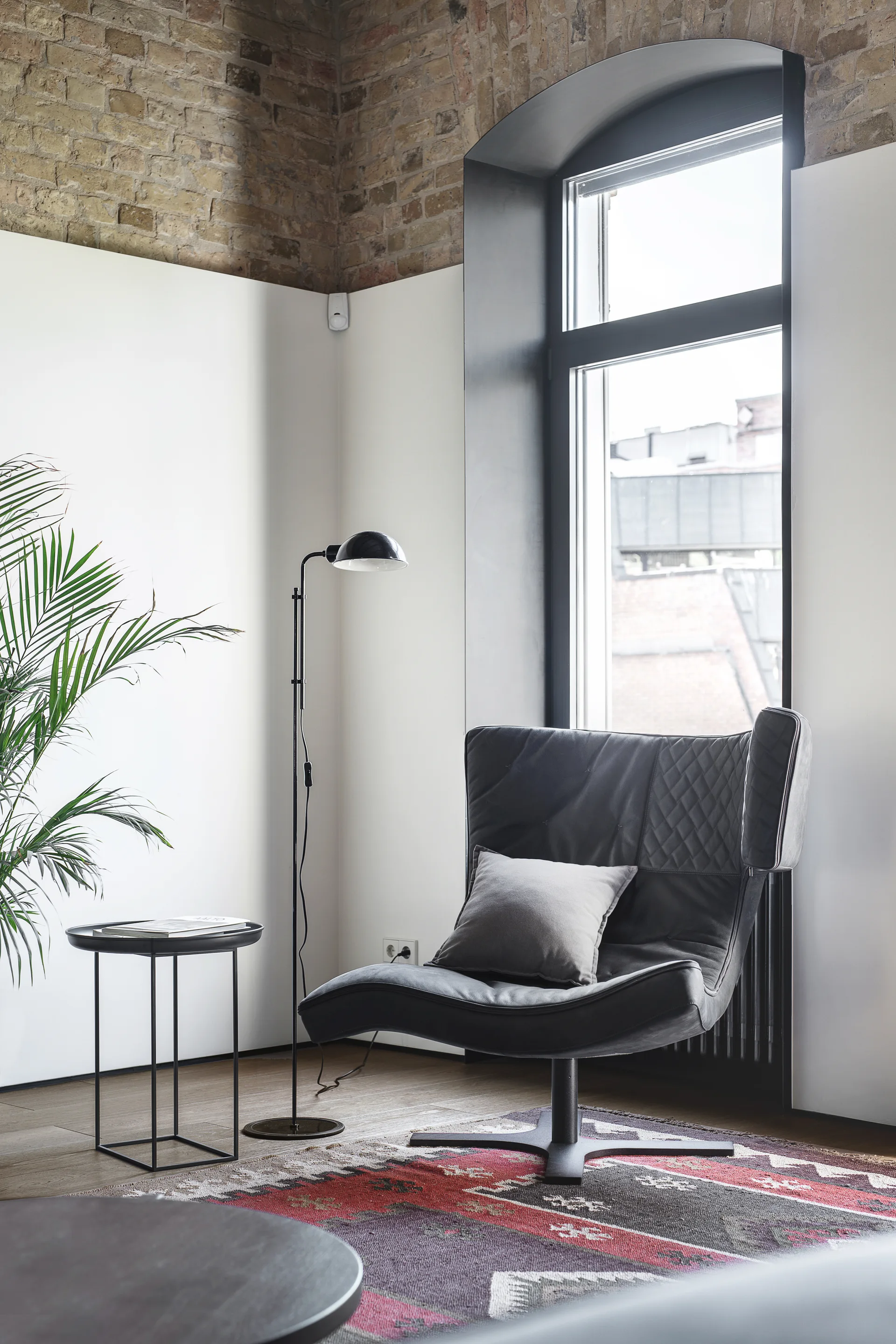 A modern reading corner features a black quilted leather armchair, a slim floor lamp, and a patterned rug beneath an arched window and exposed brick wall.