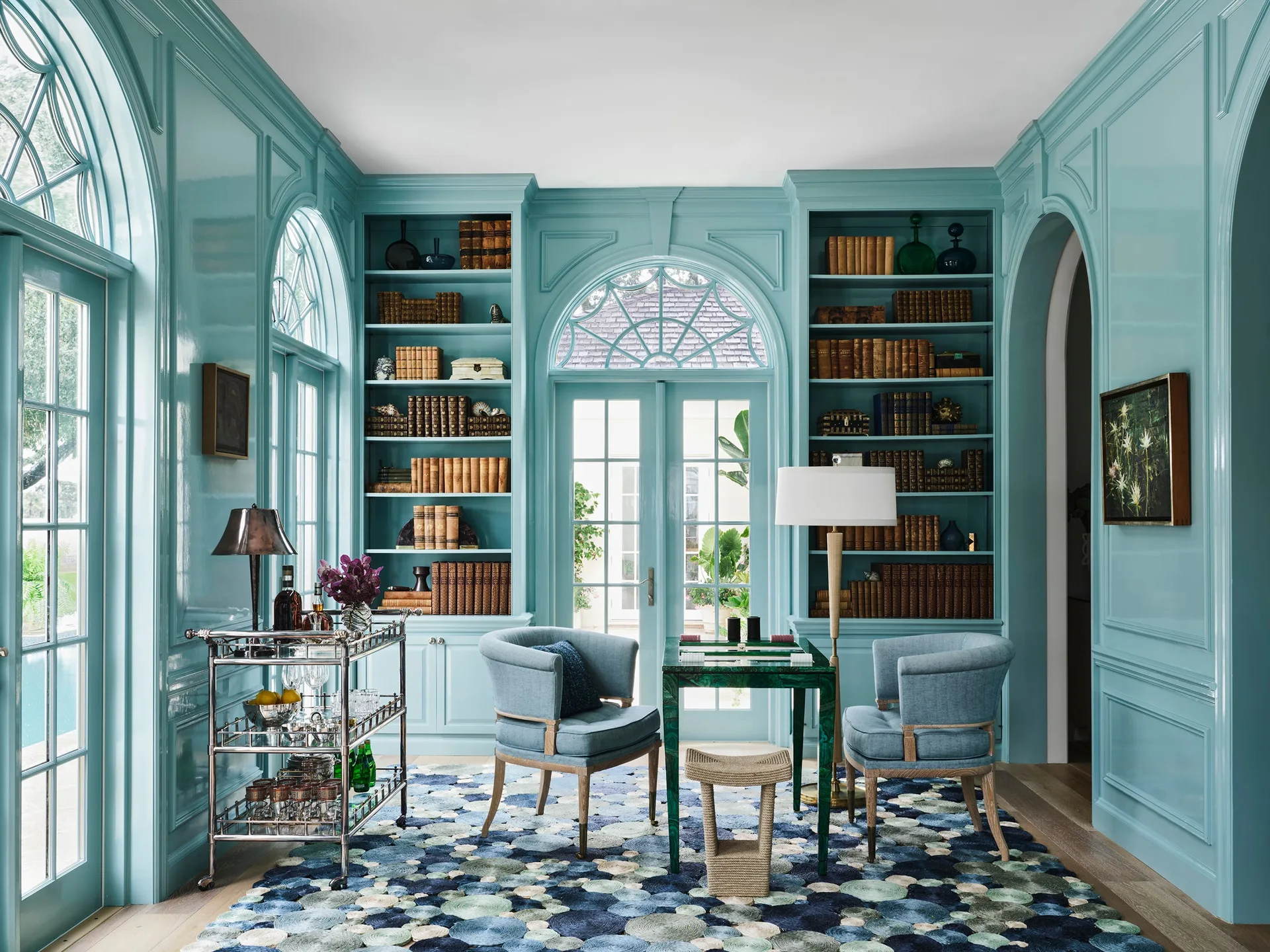 A turquoise library features high-gloss lacquered walls, matching built-in bookshelves, blue upholstered armchairs, and a green game table.