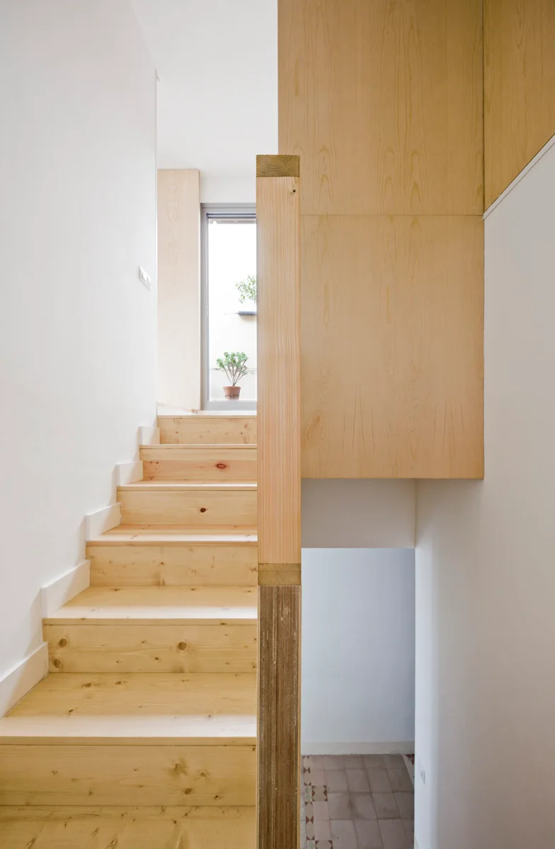 A light wood staircase with knots leads upward past white walls and a large wooden wall panel toward a window at the top.