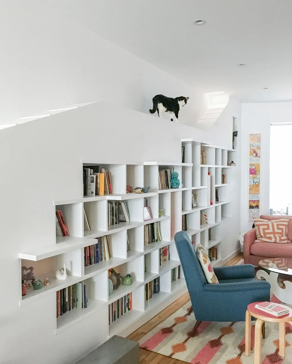 A bright living room features a custom white stepped bookcase integrated into the wall, a blue armchair, and a geometric patterned area rug.
