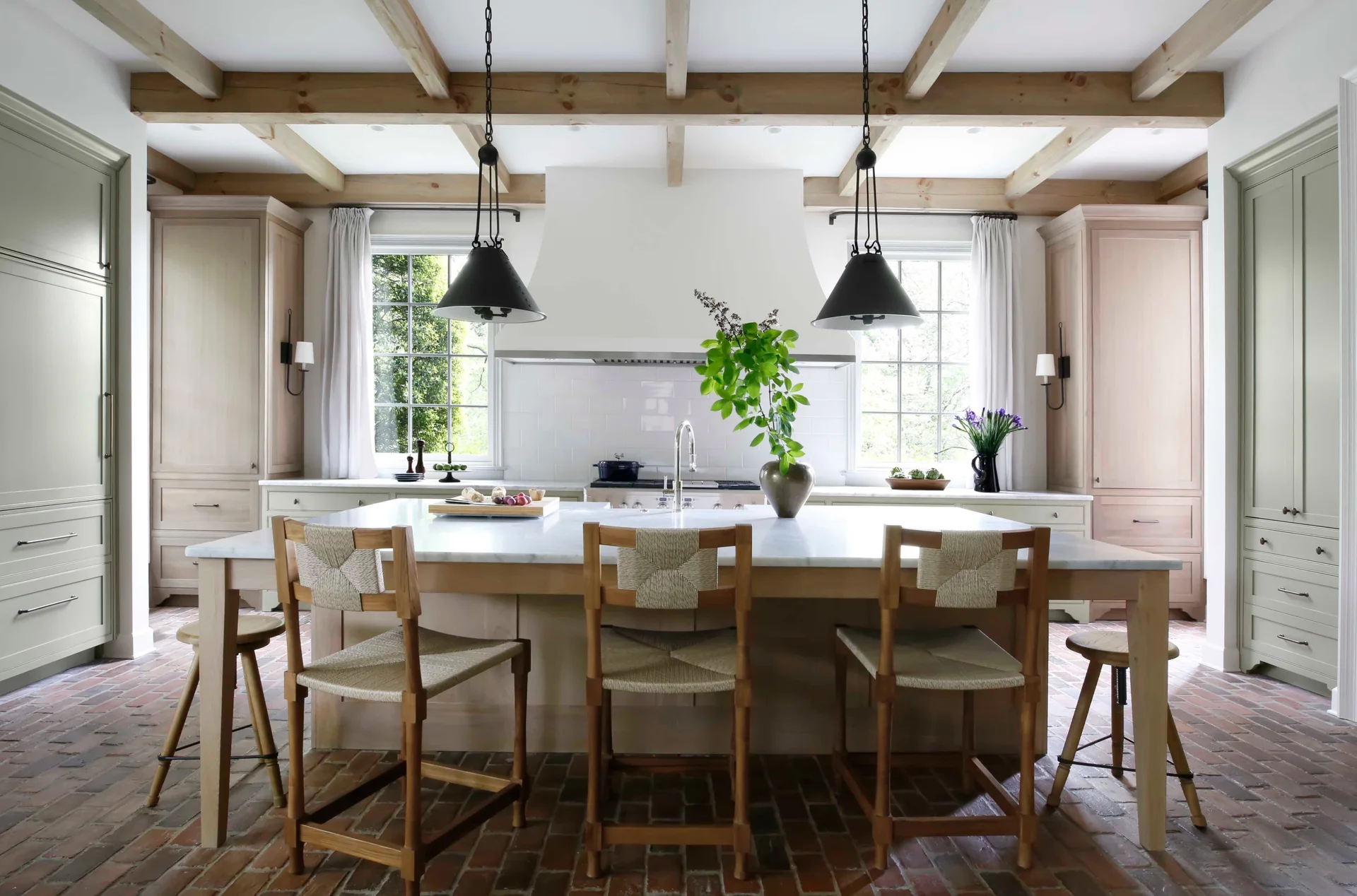 A kitchen featuring a marble-topped island with woven bar stools, sage green cabinets, exposed wood ceiling beams, and a herringbone brick floor.
