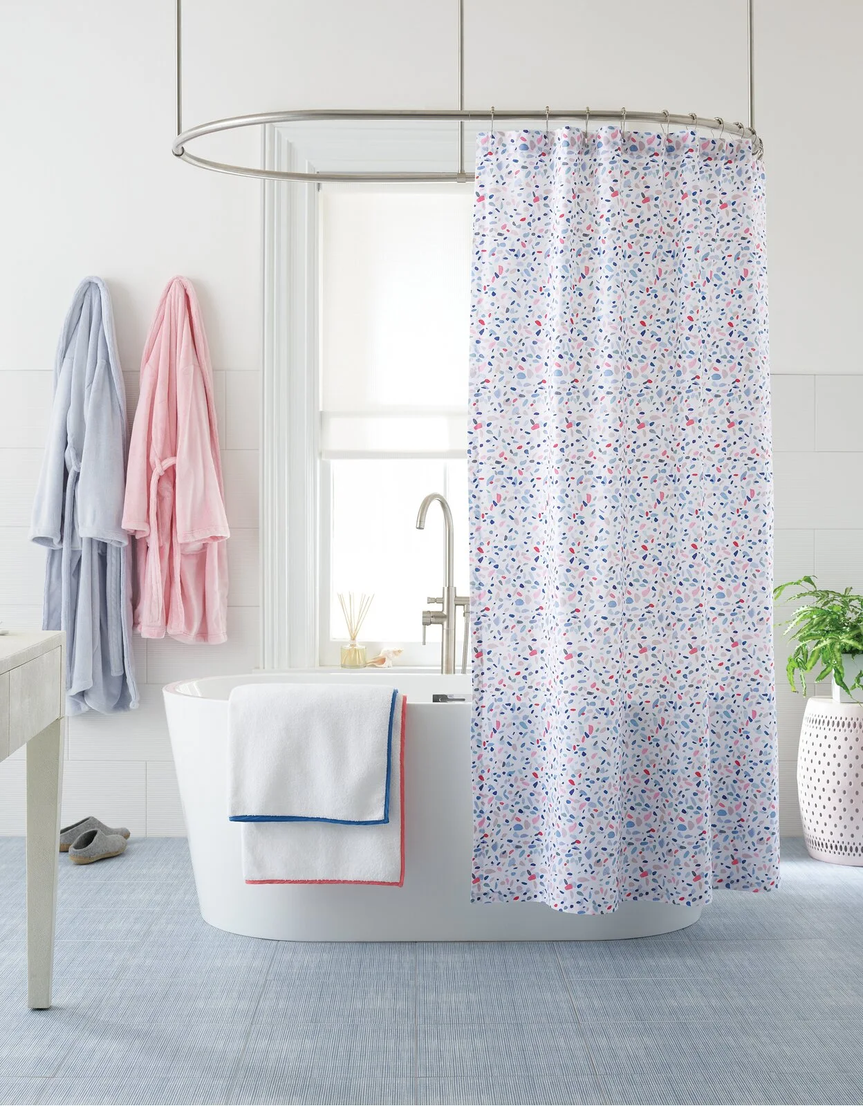 A white freestanding bathtub sits on blue tiled floor in front of a window, styled with a colorful speckled shower curtain and pastel bathrobes.