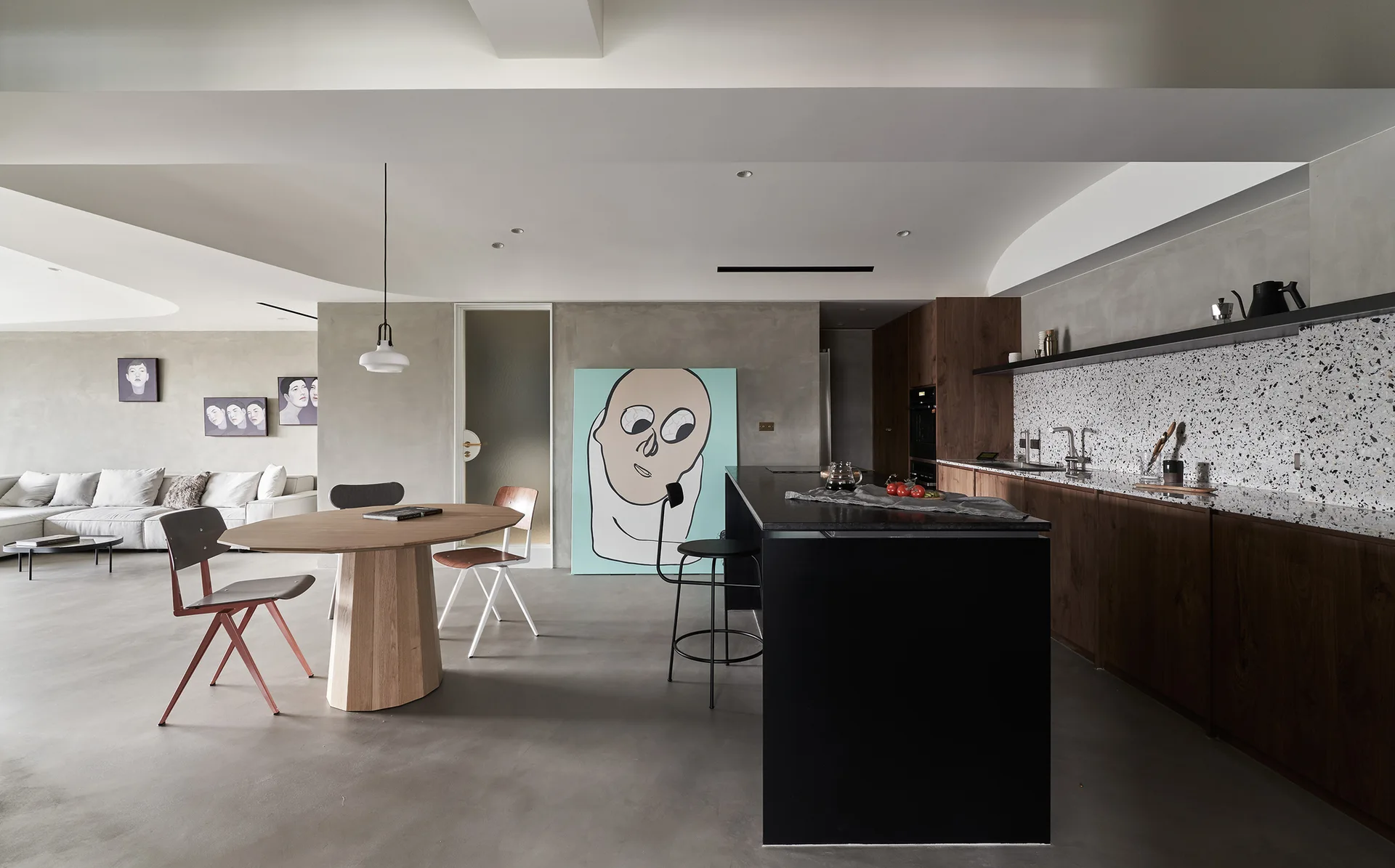 An open-concept kitchen with concrete walls, a black island, a speckled terrazzo backsplash, and an oval wooden dining table with chairs.
