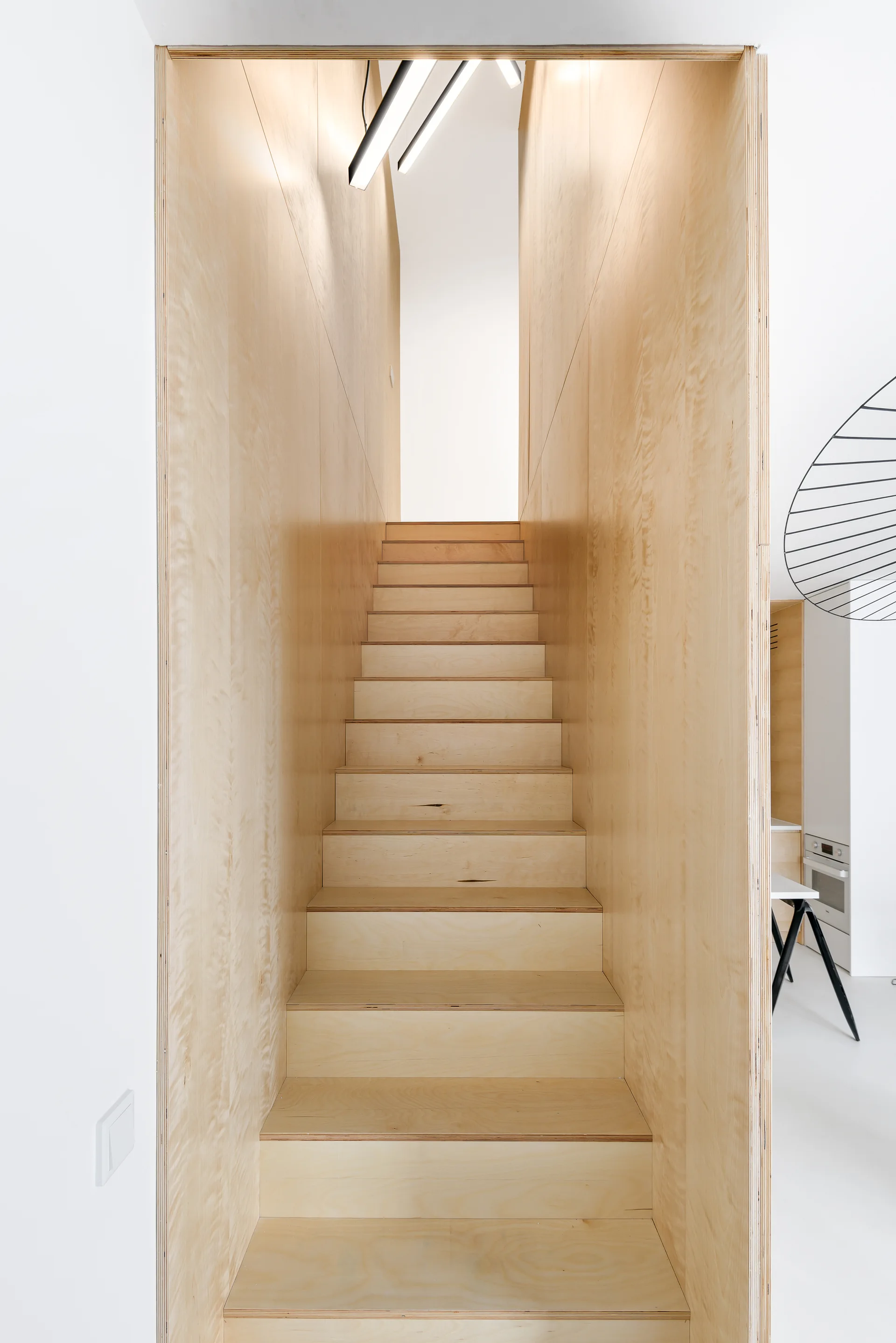 A minimalist staircase crafted from light-toned plywood steps and matching wood wall panels, set against stark white walls.