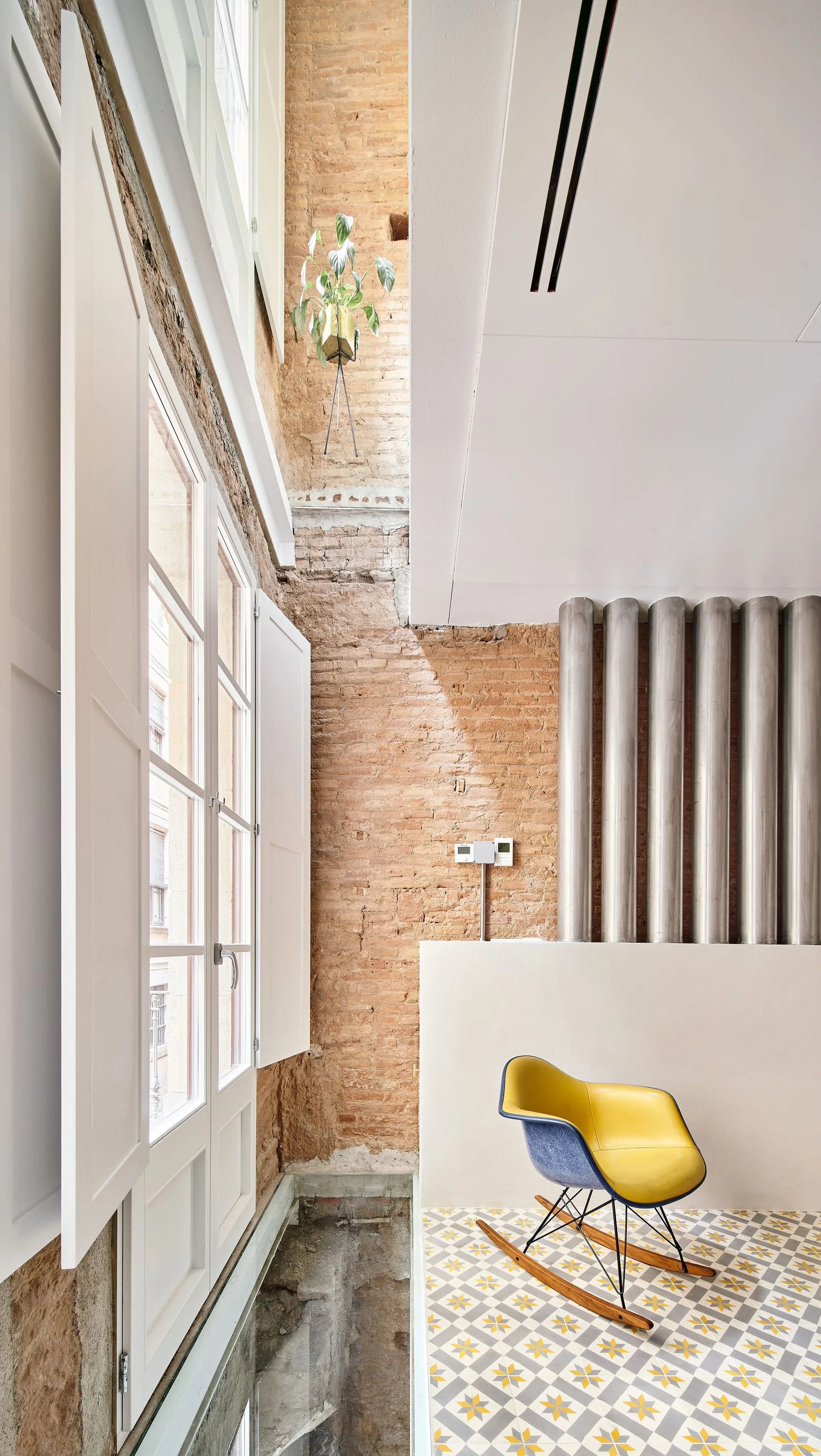 A mid-century modern rocking chair sits on yellow patterned tile floor next to an exposed brick wall with vertical metal pipes and white window shutters.