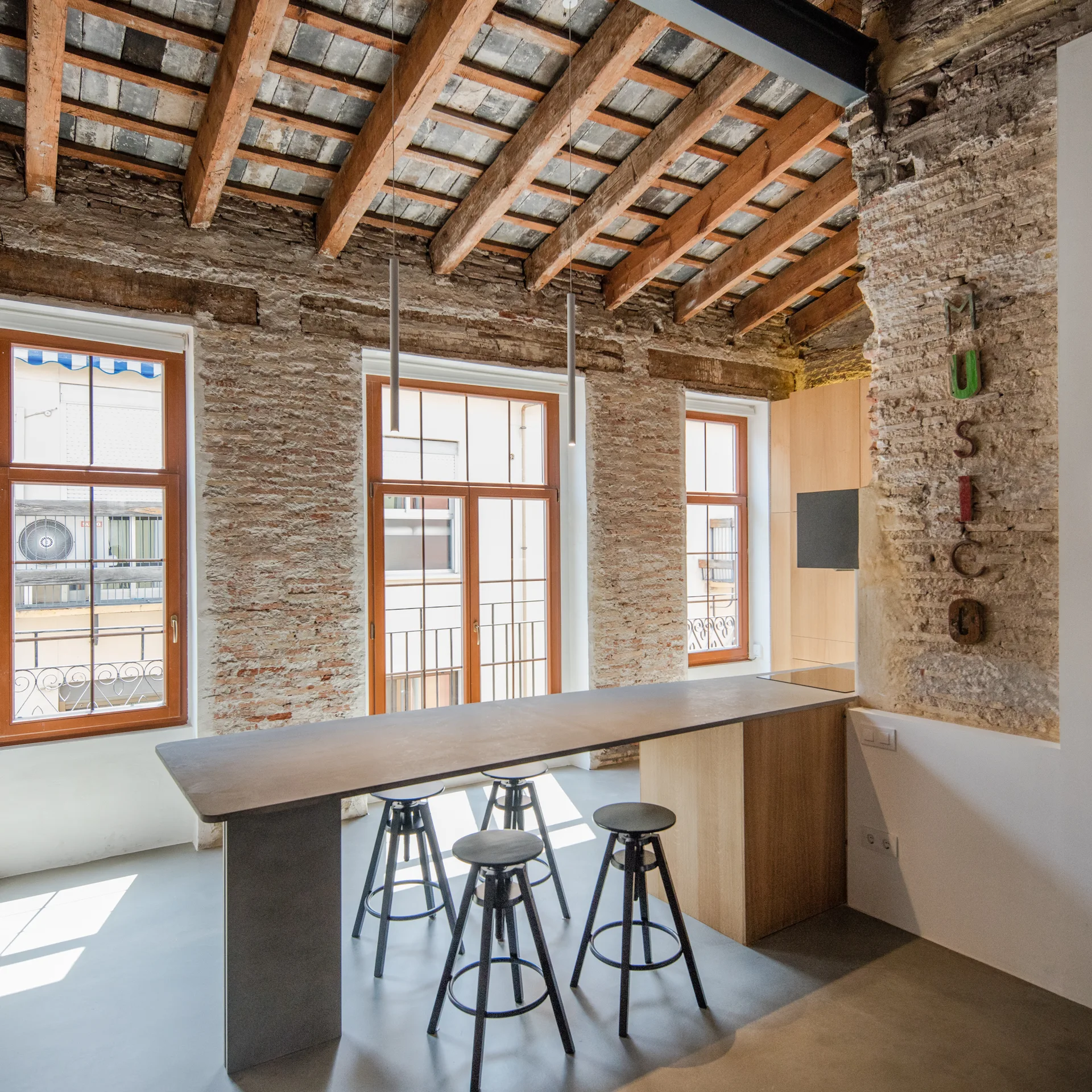 An industrial kitchen features a sleek grey breakfast bar with black stools, set against exposed brick walls and a wooden beamed ceiling.