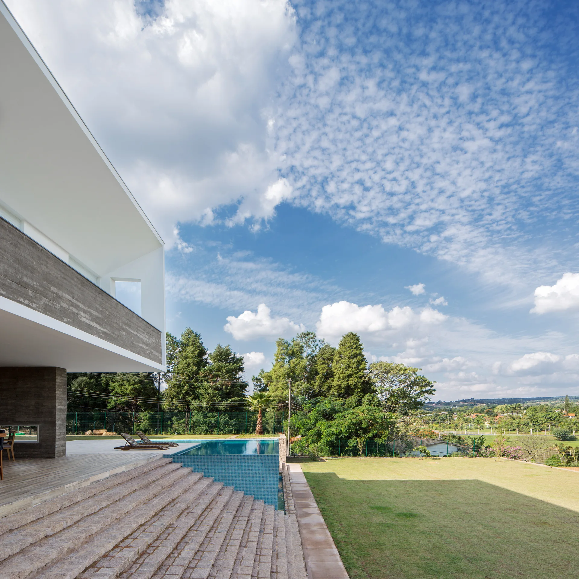 A modern residence features a concrete exterior overhang, a tiled swimming pool with lounge chairs, and wide stone steps leading down to a green lawn.