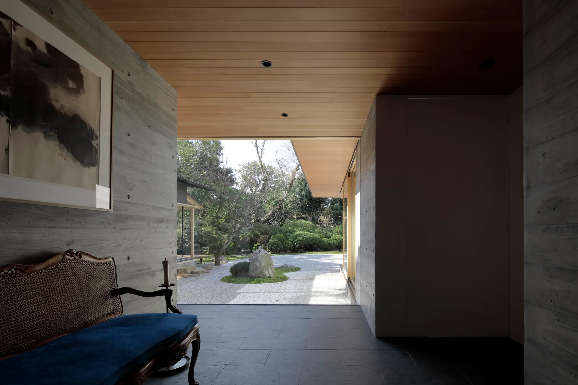 A modern concrete entryway featuring a vintage blue settee, dark slate floors, and an open doorway leading to a Japanese gravel garden.