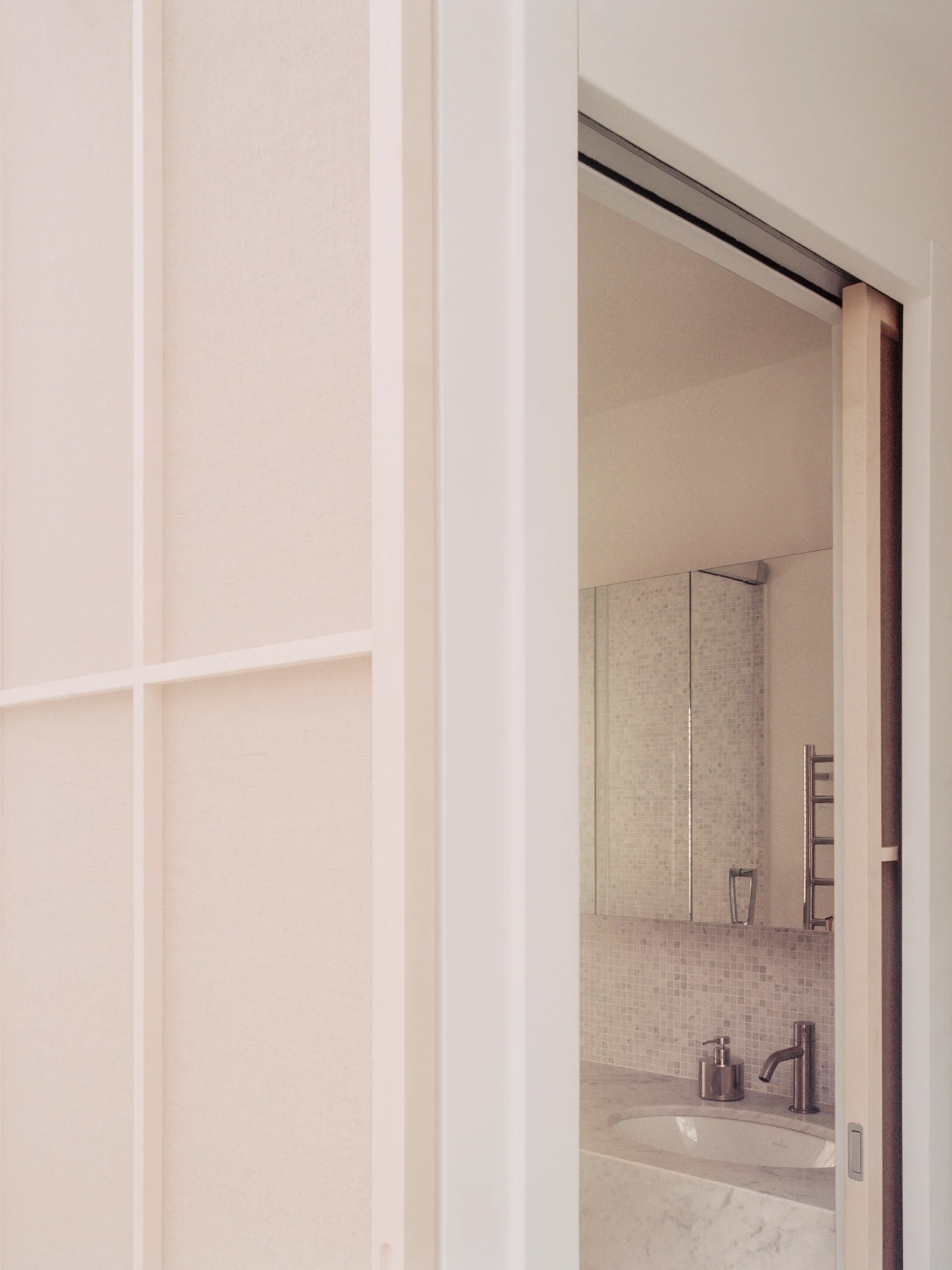 An open white grid sliding door reveals a minimalist bathroom featuring a marble vanity, mosaic tile backsplash, and mirrored cabinet.