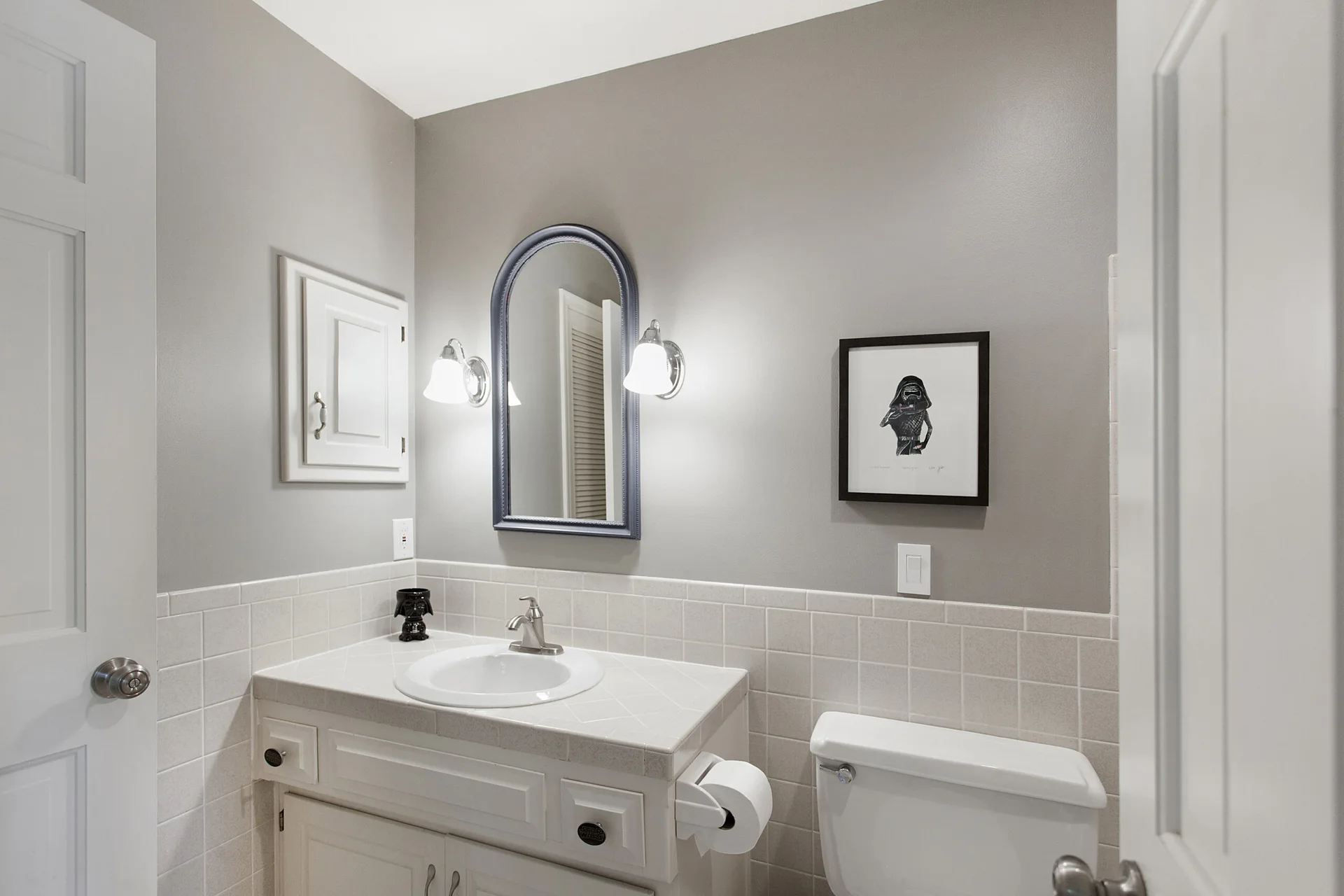 A bathroom vanity with a white tiled countertop and backsplash, an arched dark-framed mirror, and a small built-in wall cabinet.
