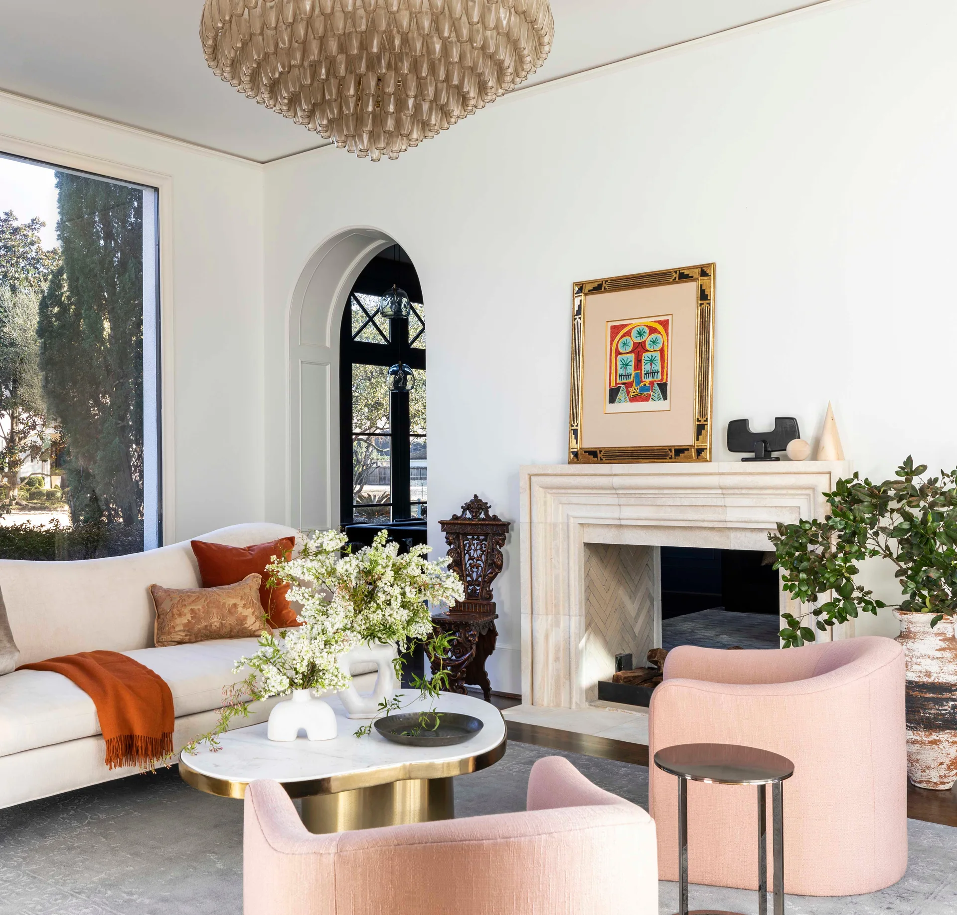 A bright living room features two pink armchairs, a cream sofa with rust pillows, an oval marble coffee table, and a carved stone fireplace.