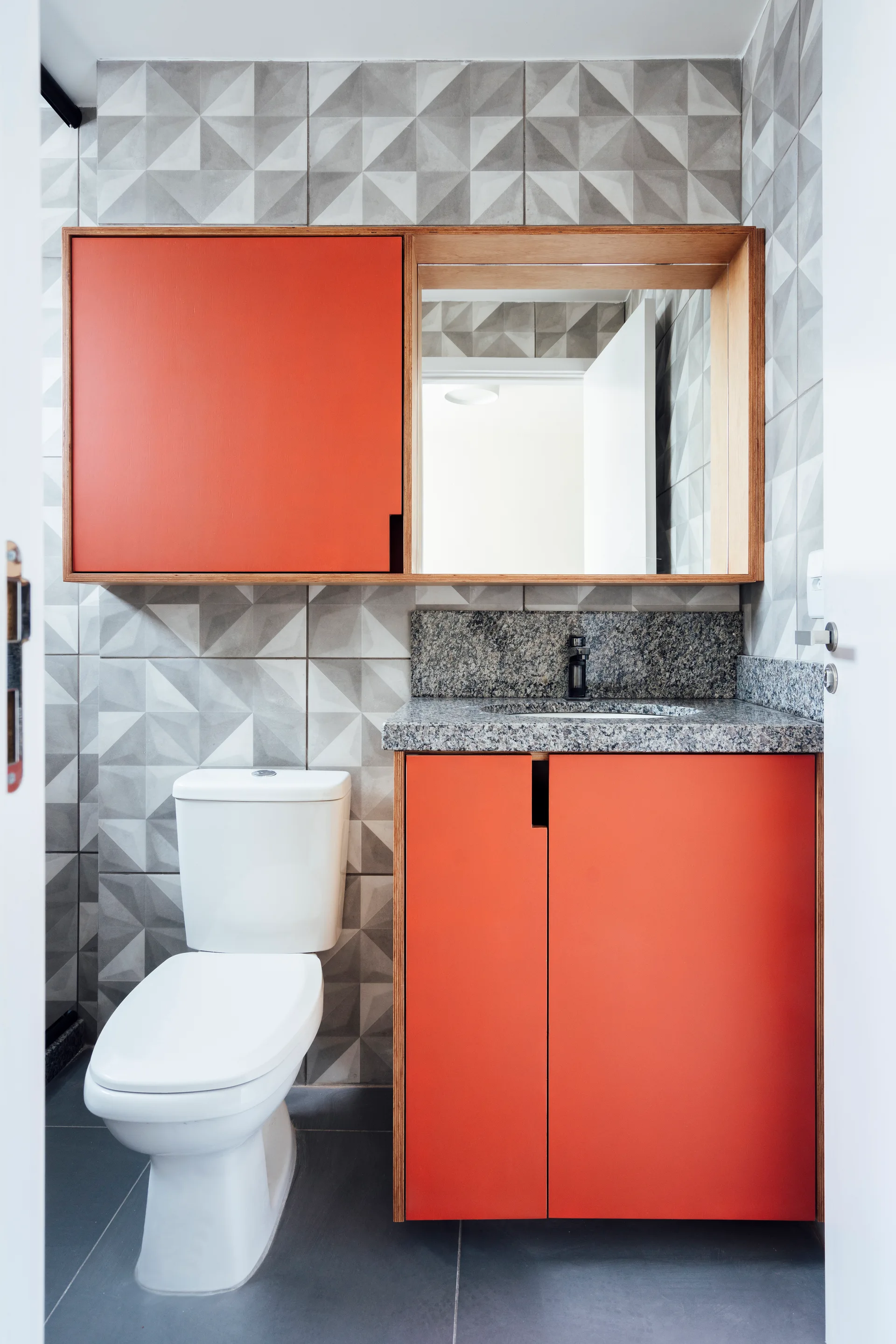 A modern bathroom vanity area features vibrant orange cabinetry, a rectangular mirror, speckled grey granite countertop, and 3D geometric wall tiles.