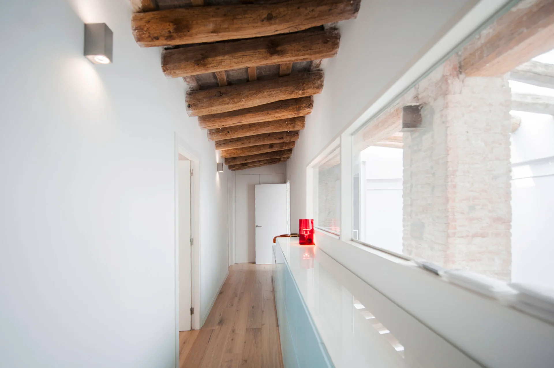 A long white hallway featuring rustic exposed log ceiling beams, a light wood floor, and a low glossy built-in cabinet beneath horizontal windows.