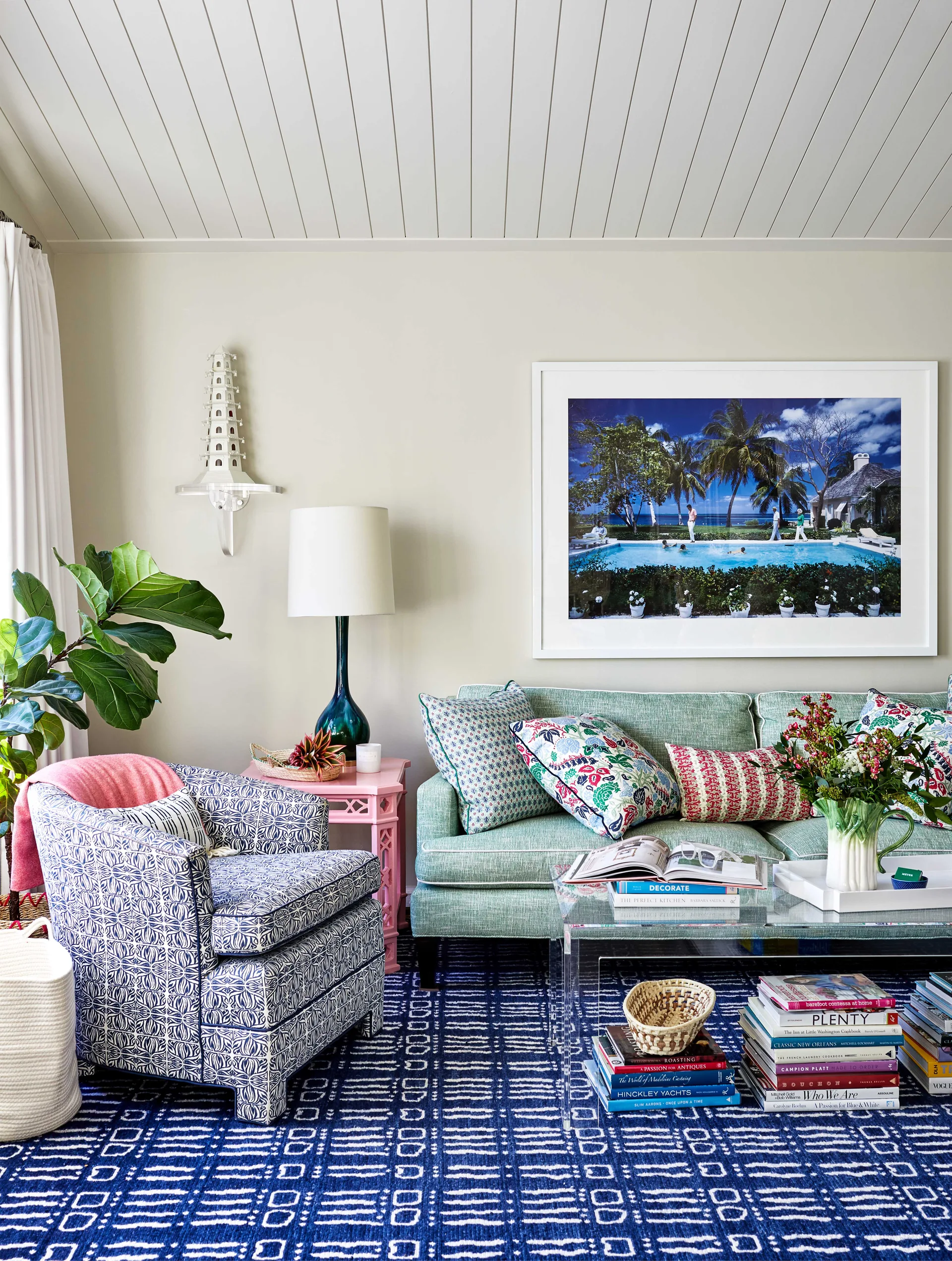 An eclectic living room features a green sofa with colorful pillows, a patterned blue armchair, and a clear acrylic coffee table over a blue patterned rug.