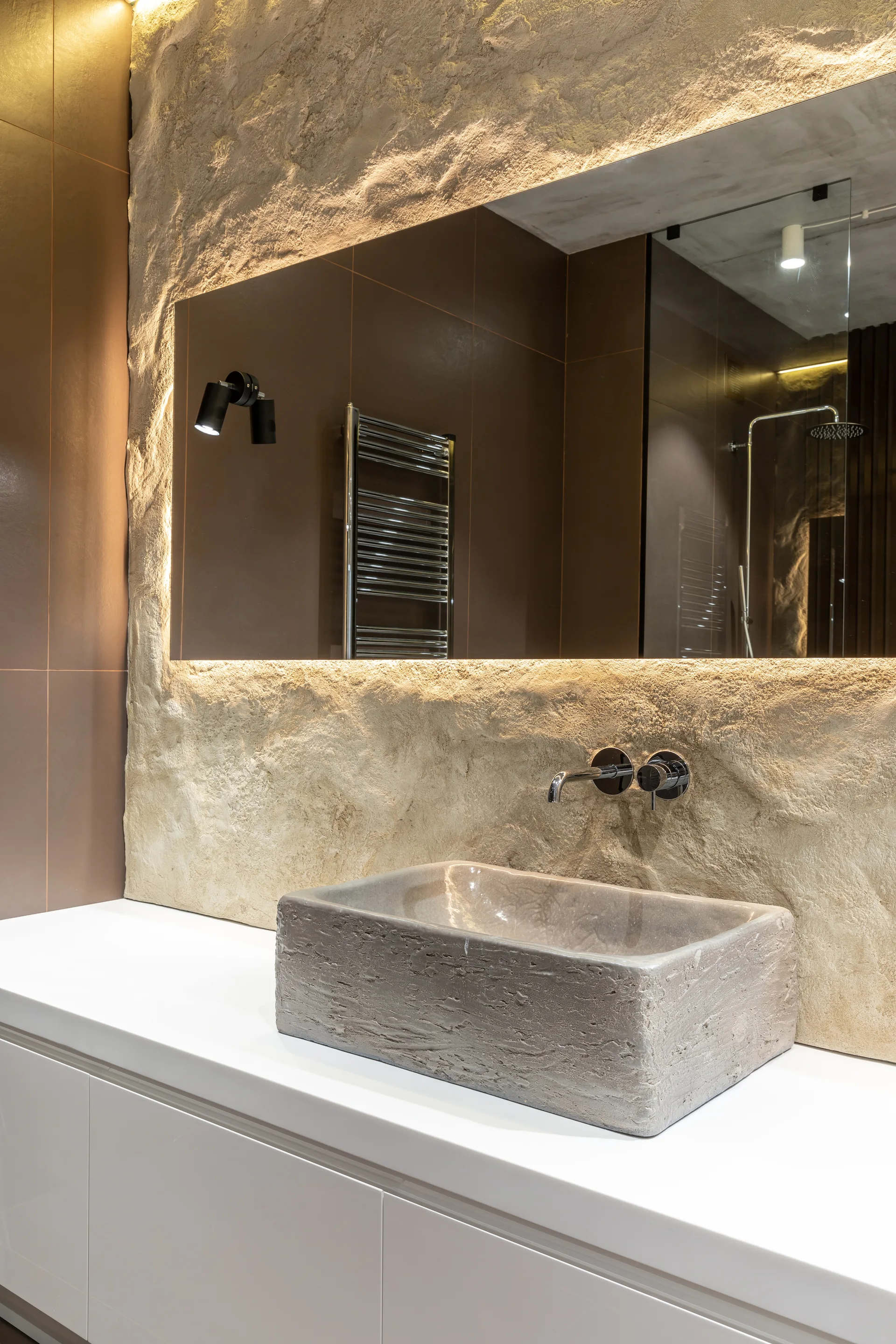 A textured stone vessel sink sits on a white vanity countertop against a rough beige rock wall with a backlit mirror and chrome faucet.