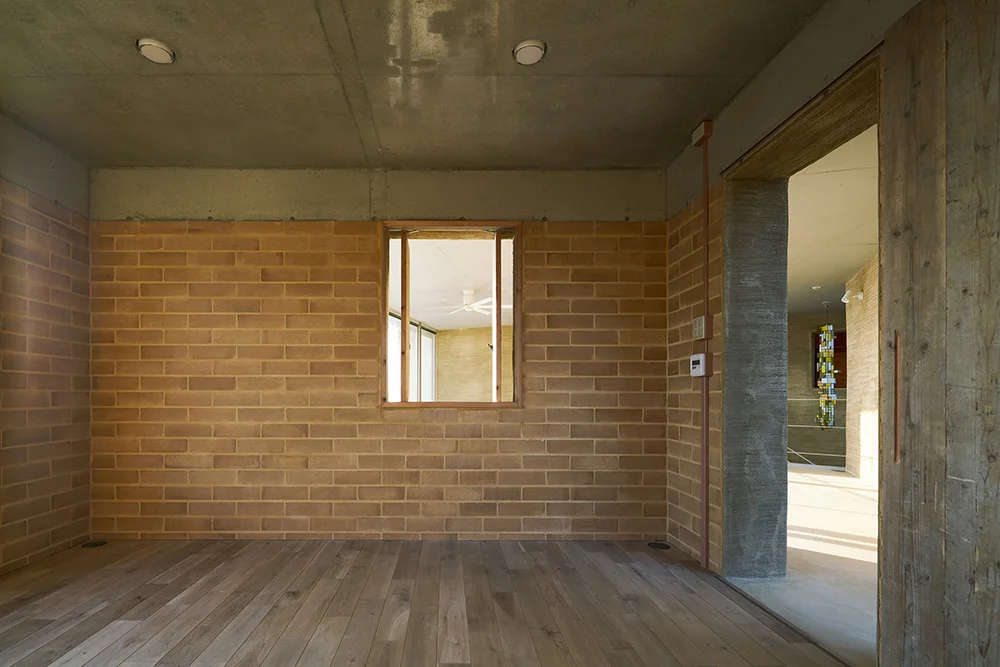 An empty room featuring exposed light brown brick walls, a raw concrete ceiling, wood plank flooring, and an interior window.