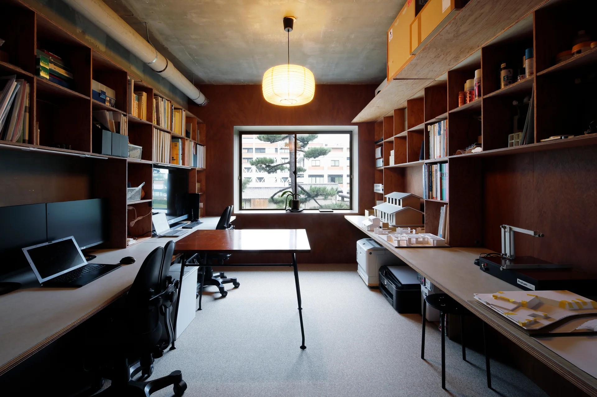 A home office with custom plywood shelving, built-in desks, black office chairs, a paper lantern pendant light, and an exposed concrete ceiling.