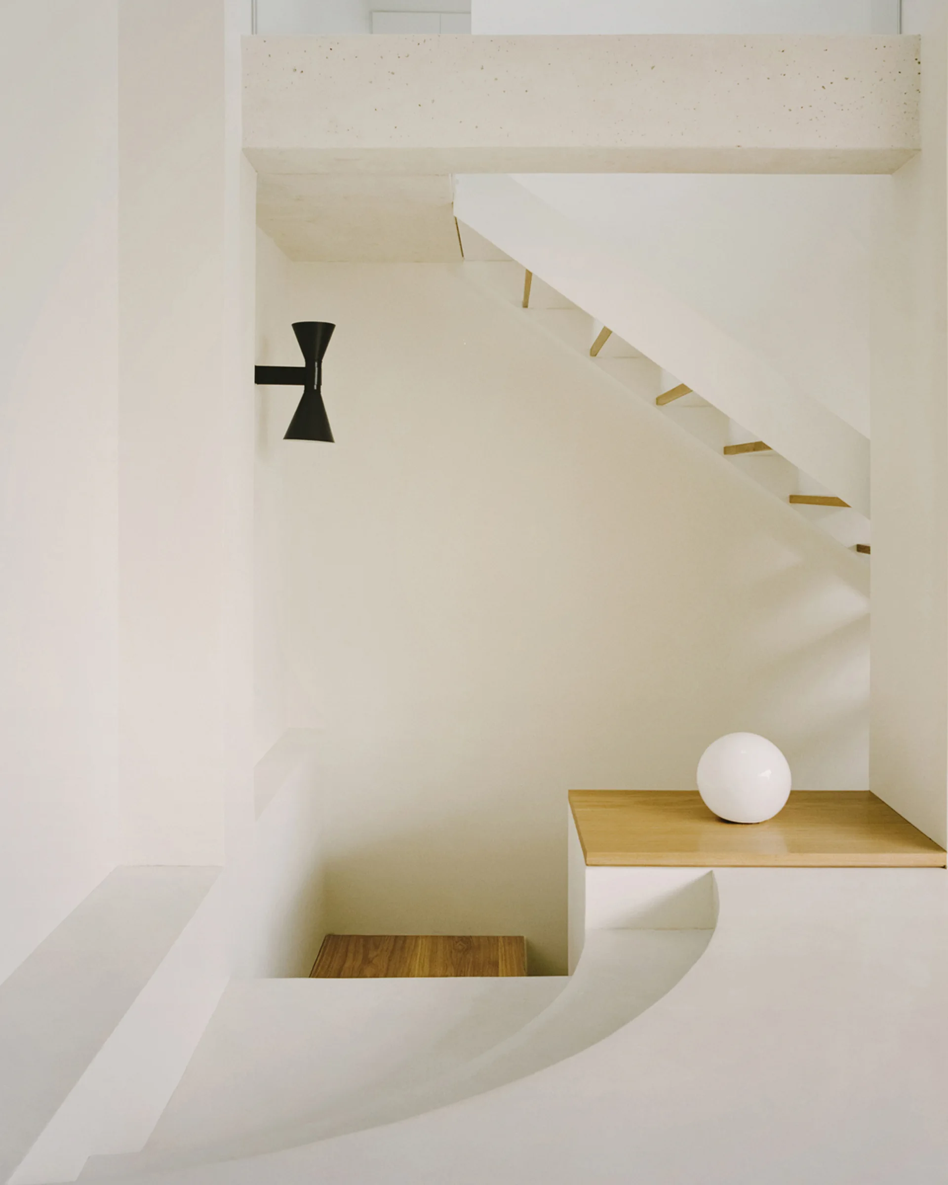 A minimalist white stairwell featuring smooth plaster walls, a black hourglass wall sconce, a wooden ledge with a white globe lamp, and a modern staircase.