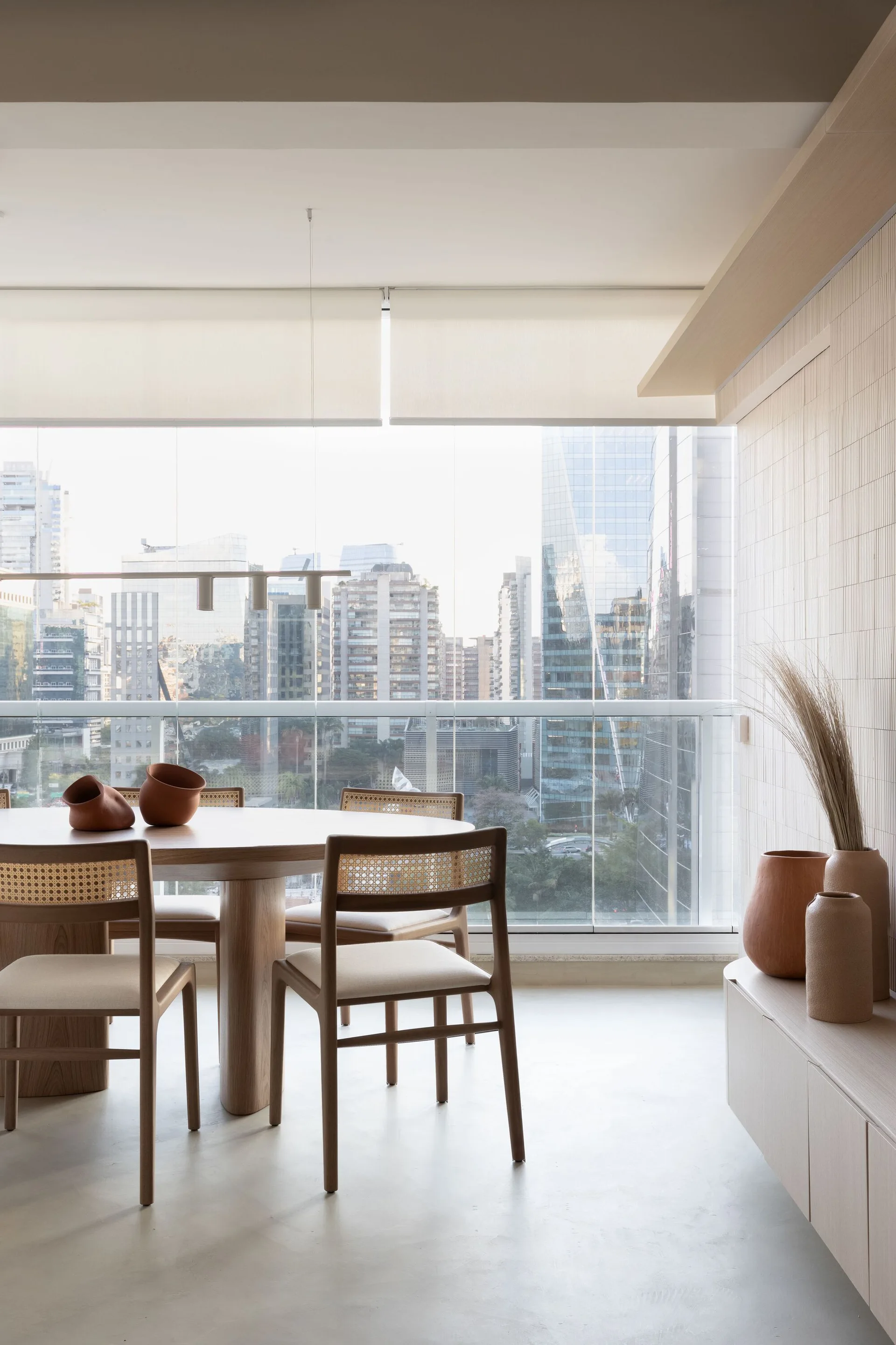 A minimalist dining room features a round light wood table and cane-back chairs next to a large window with city views and a slatted accent wall.