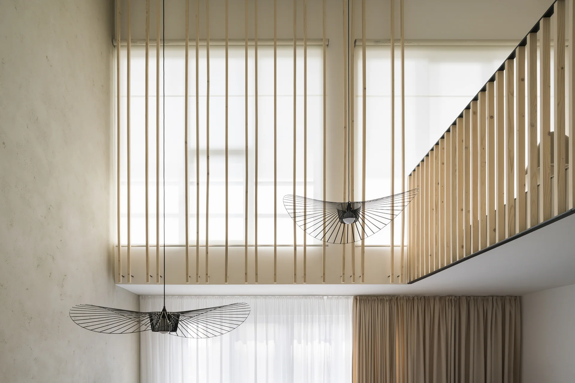 Double-height loft interior featuring a light wood slatted screen over the window, matching mezzanine railing, and two black sculptural pendant lights.