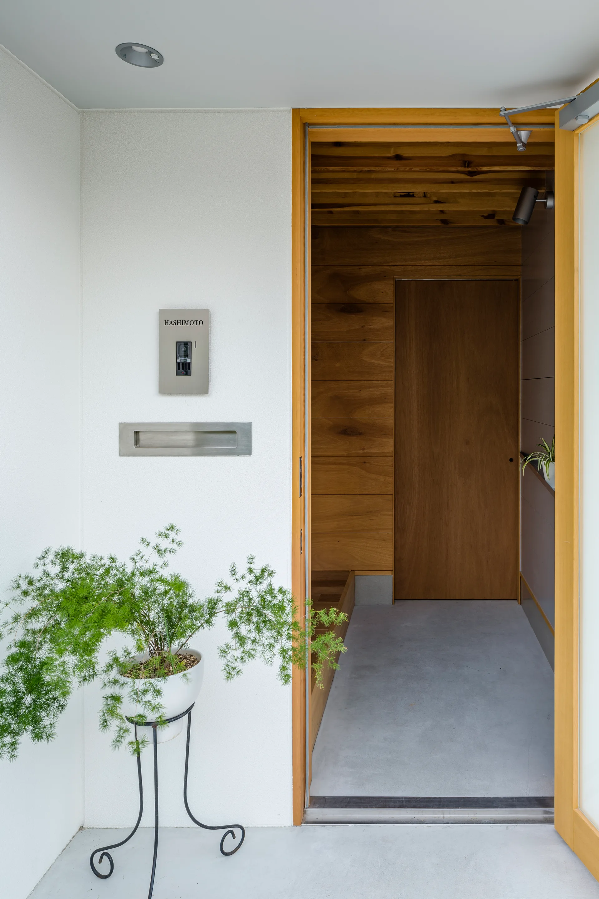 A modern home entryway features a white exterior wall, a natural wood door frame, and a warm wood-paneled interior hallway.