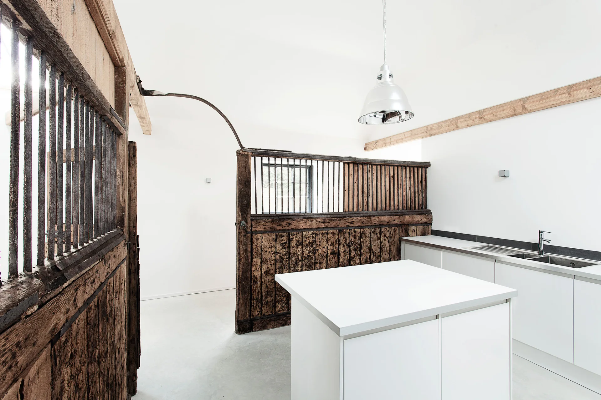 A modern white kitchen features a large white island and a rustic, weathered wood horse stall partition as a room divider.