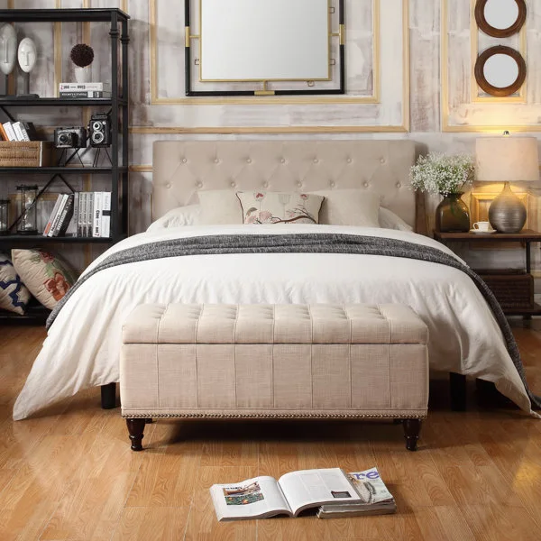 A bedroom features a beige tufted headboard and matching bench, a black metal bookshelf, and a large wall mirror on paneled walls.
