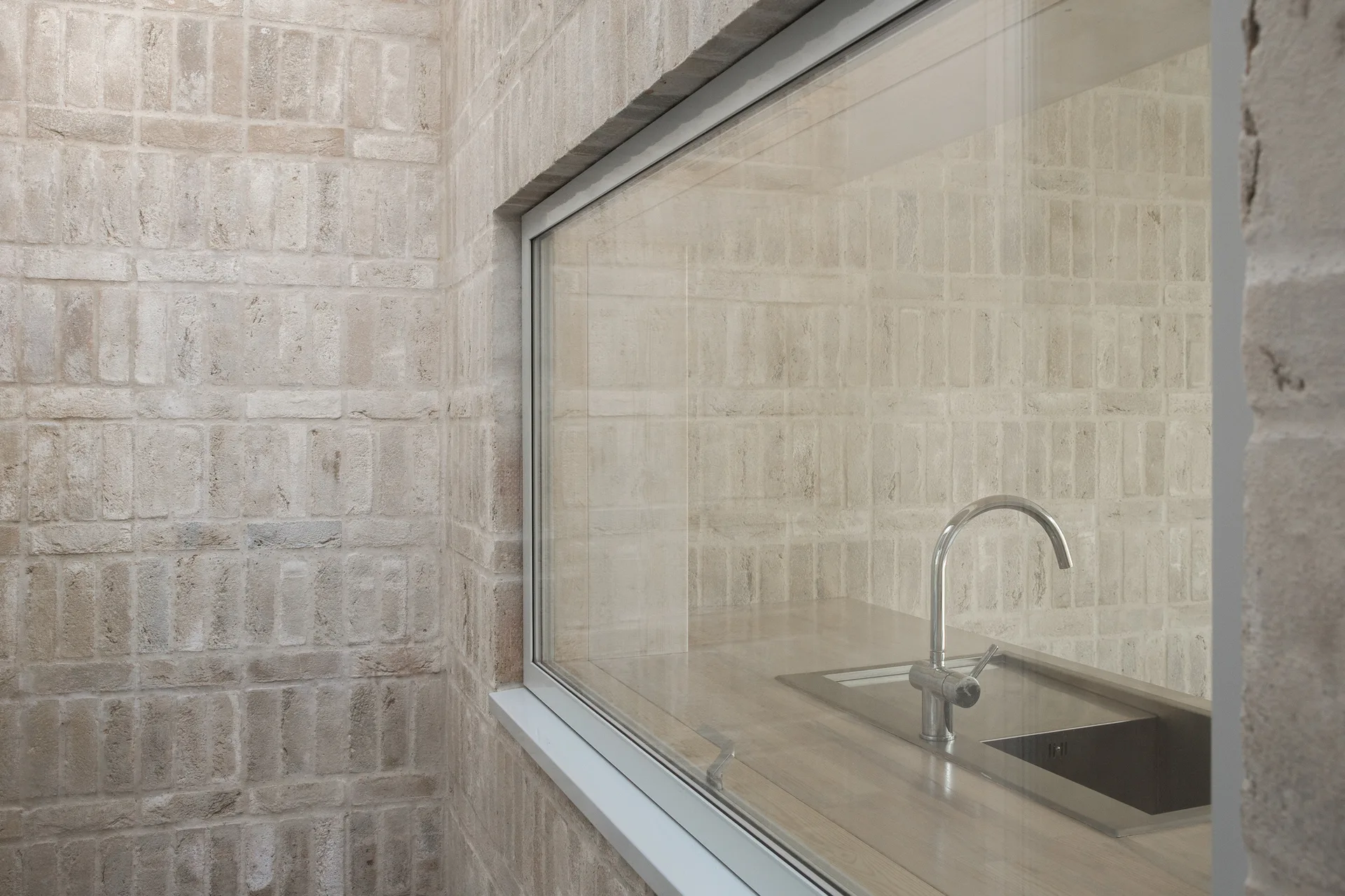 A chrome faucet and sink on a wood countertop are visible through a large glass window set in a light beige brick wall.