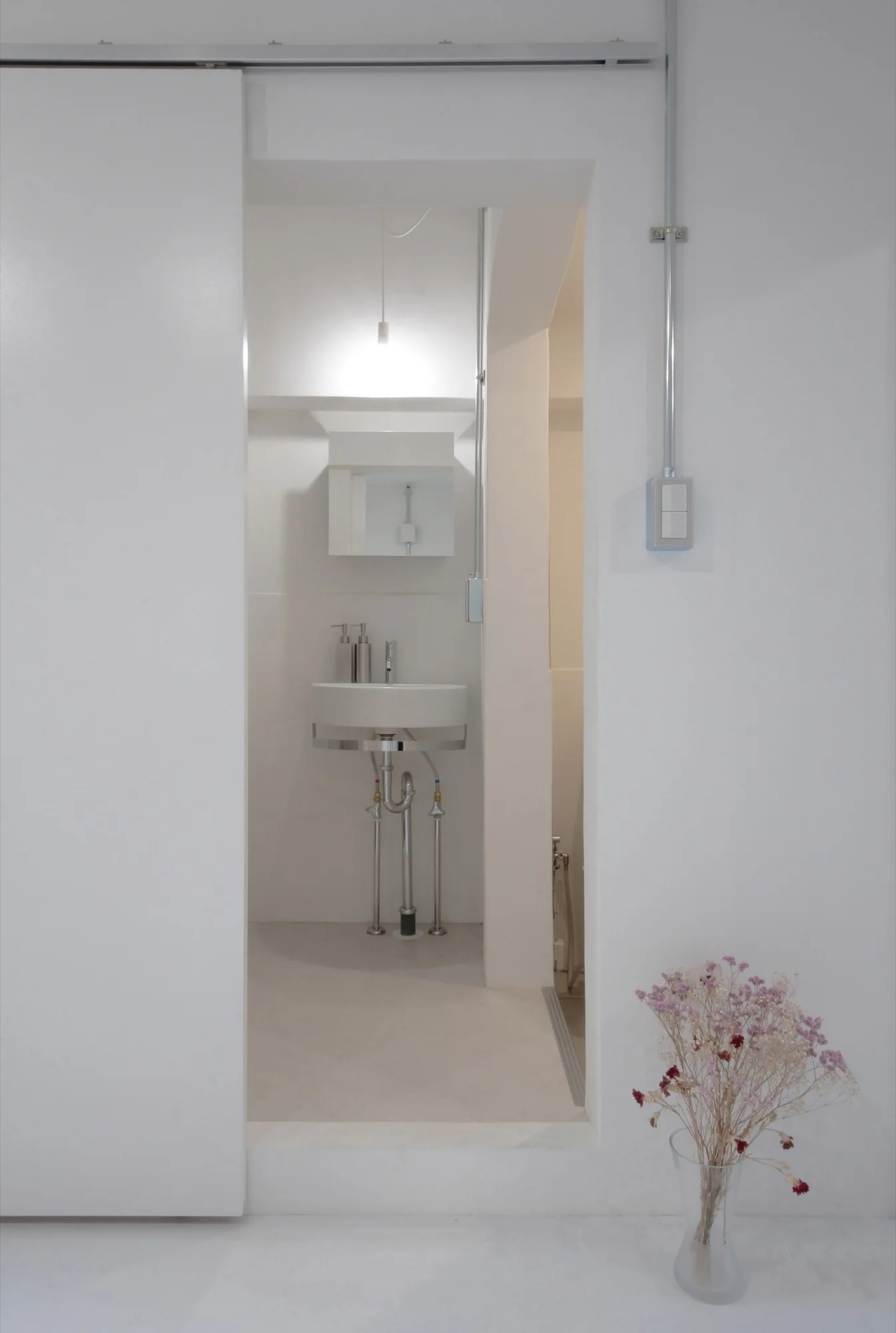 A minimalist white powder room featuring a wall-mounted sink with exposed chrome plumbing, a mirror, and an exposed metal conduit on the wall.