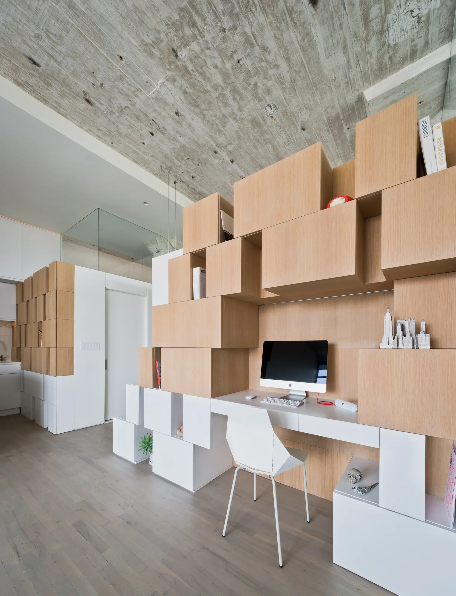 A modern home office workspace featuring a custom staggered wood block shelving unit, an integrated white desk, and an exposed concrete ceiling.