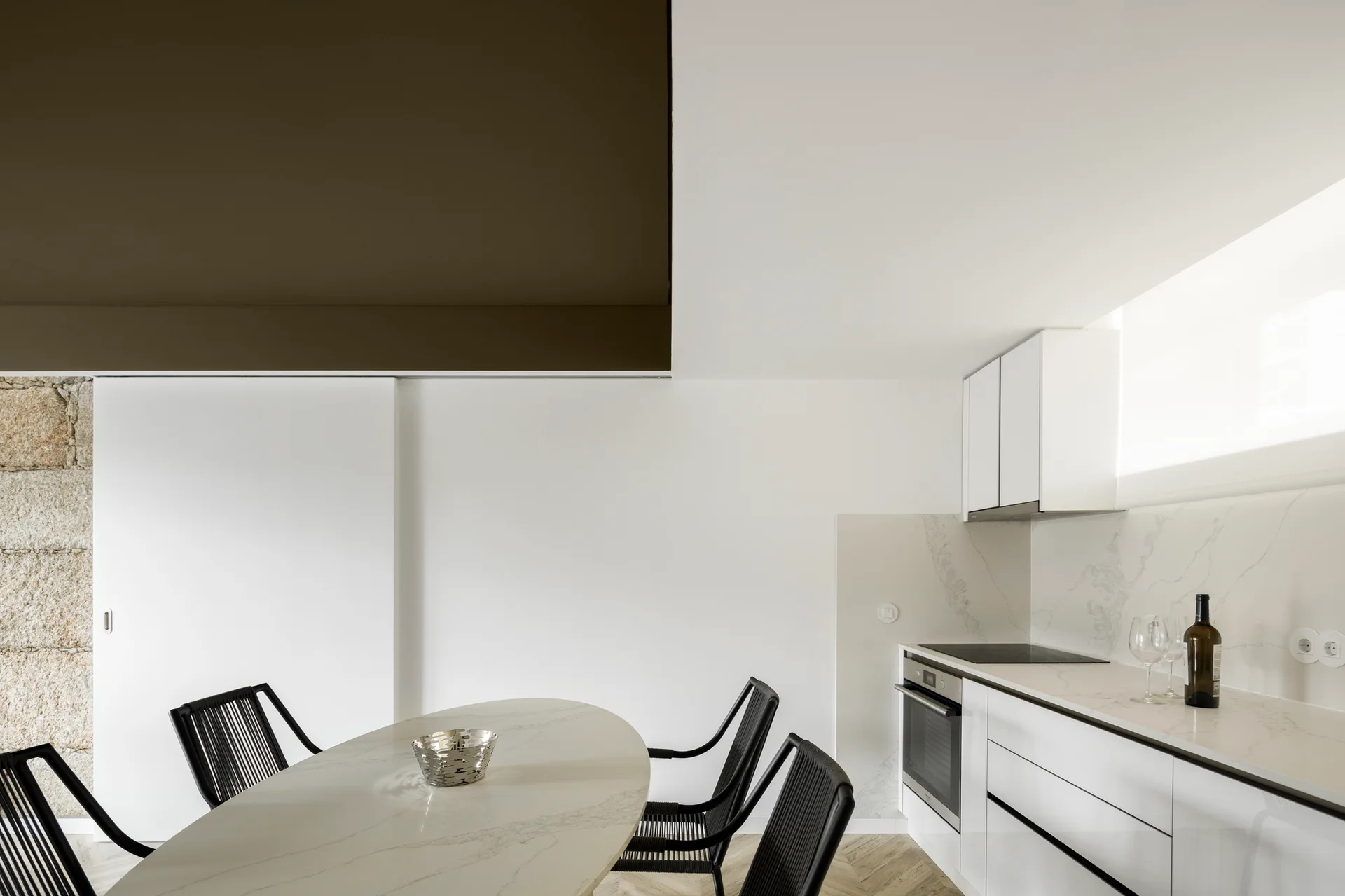 A minimalist white kitchen features an oval marble-topped dining table with black woven chairs, high-gloss cabinetry, and an exposed stone wall.
