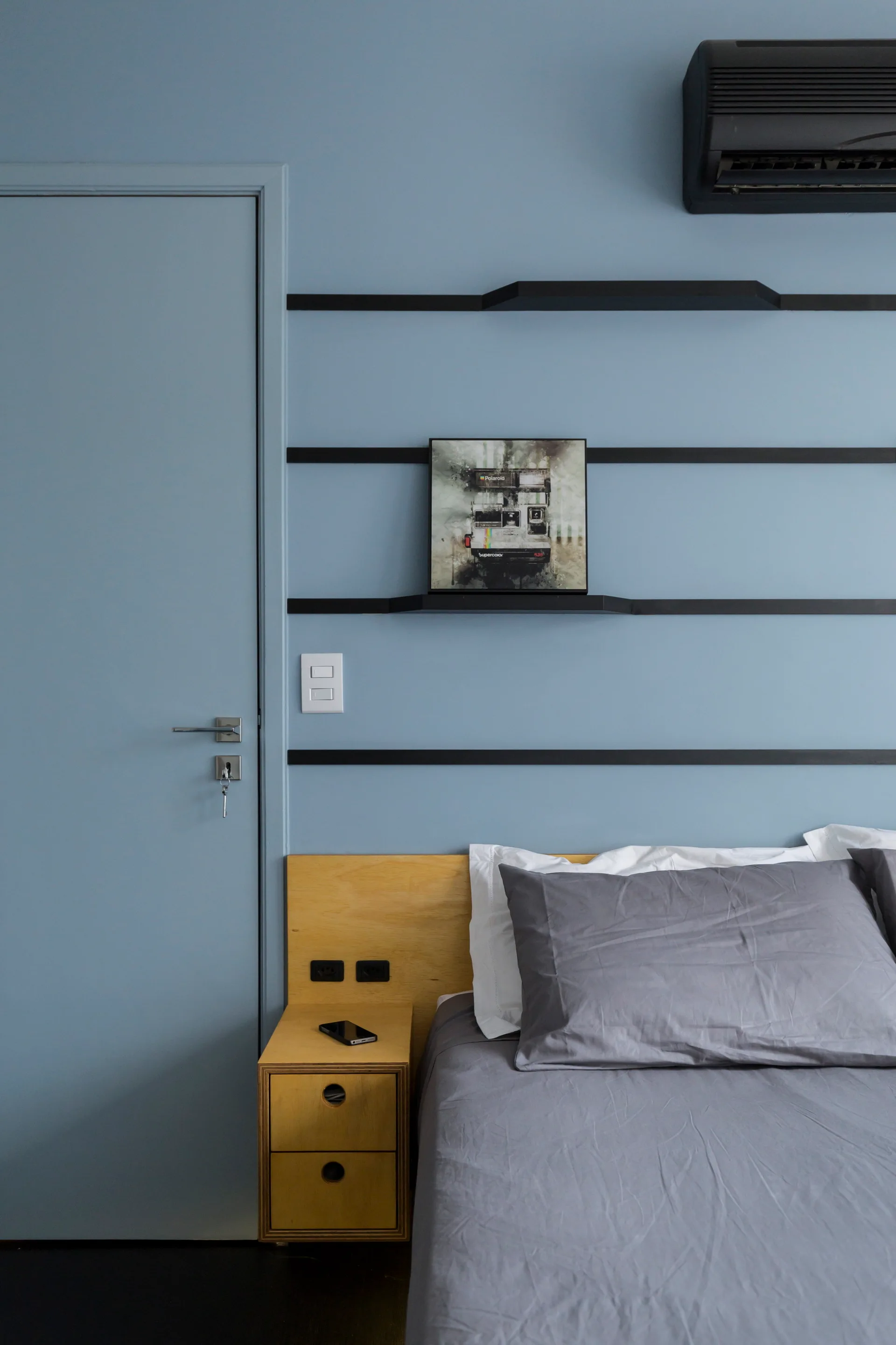 A blue bedroom wall with black horizontal shelves features an integrated light wood nightstand and bed with grey linens.