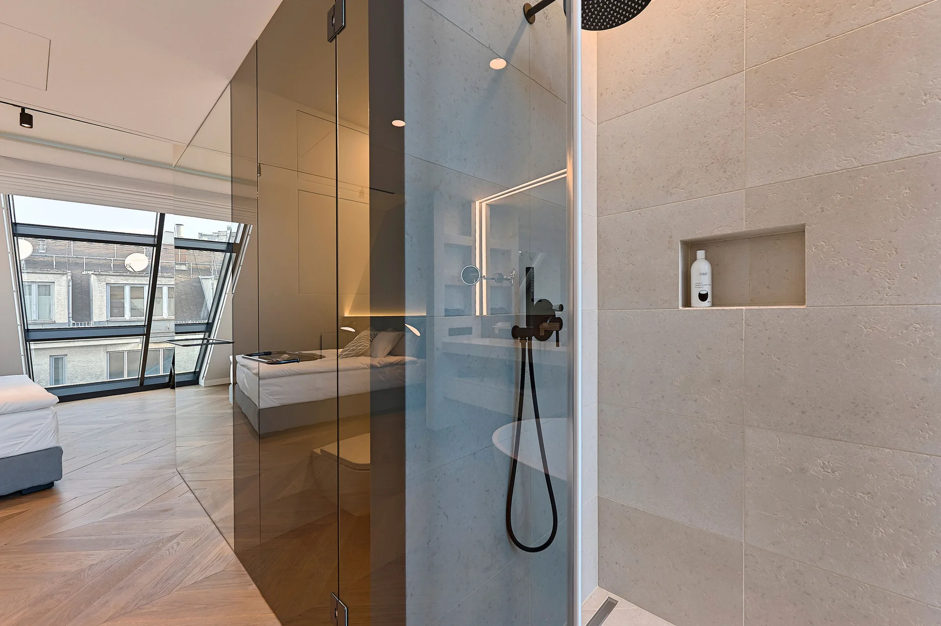 A walk-in shower with beige stone-look tile walls, matte black fixtures, a recessed niche, and a glass partition reflecting an adjacent bedroom.