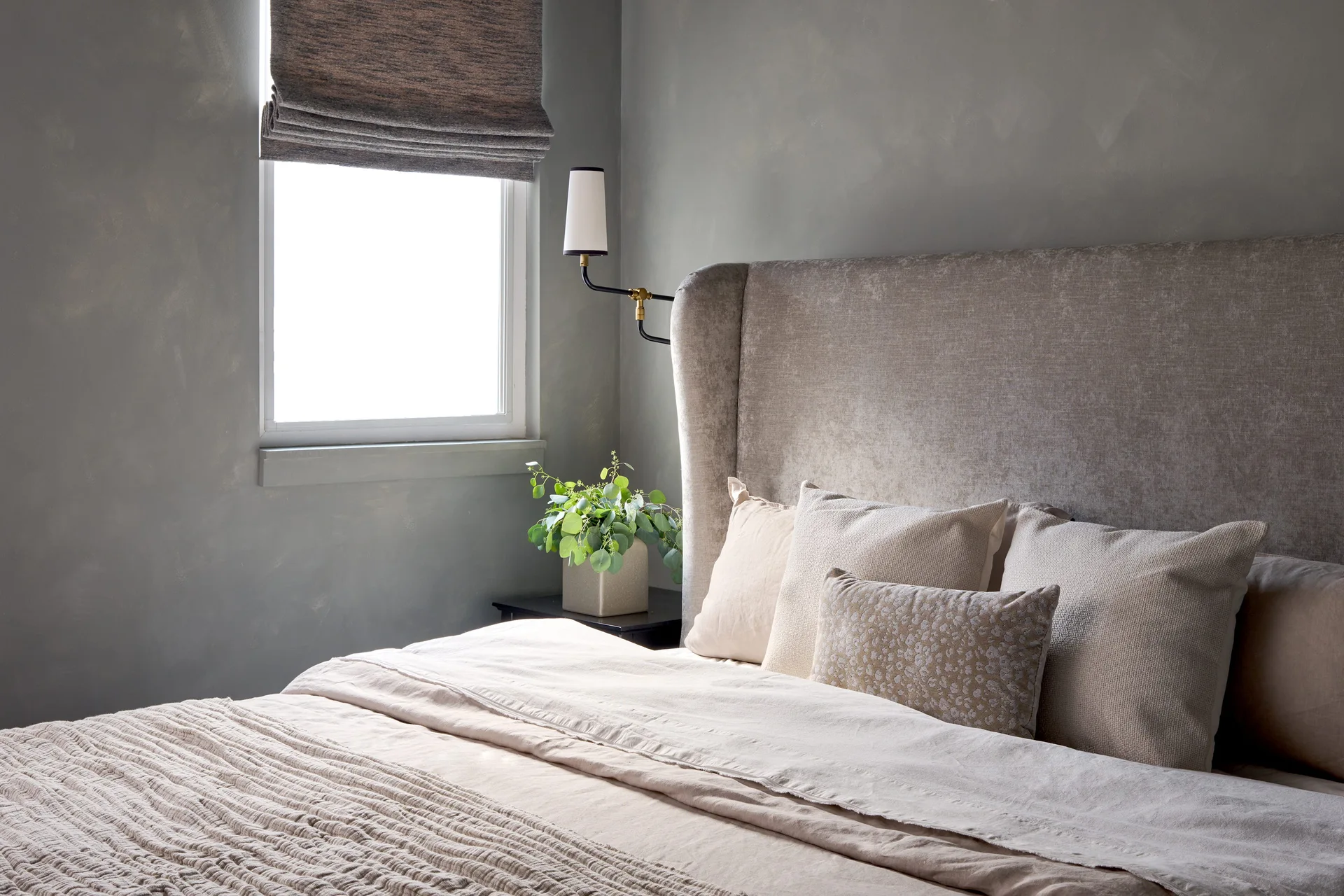 A neutral bedroom features a textured upholstered headboard, a black wall sconce, and a window with a fabric roman shade next to a potted plant.