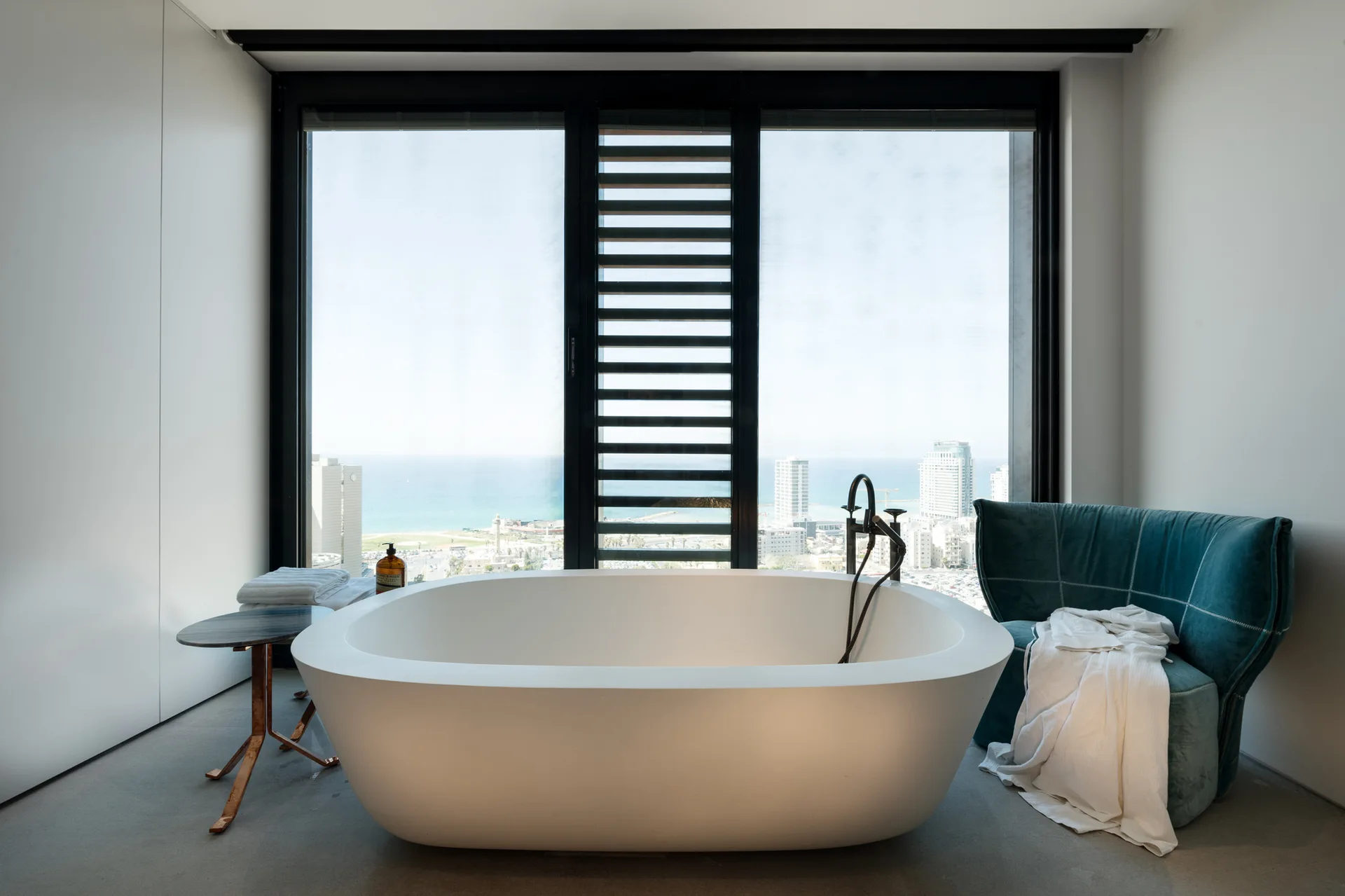 A white freestanding bathtub stands before a large window overlooking a coastal city, flanked by a small side table and a teal armchair.