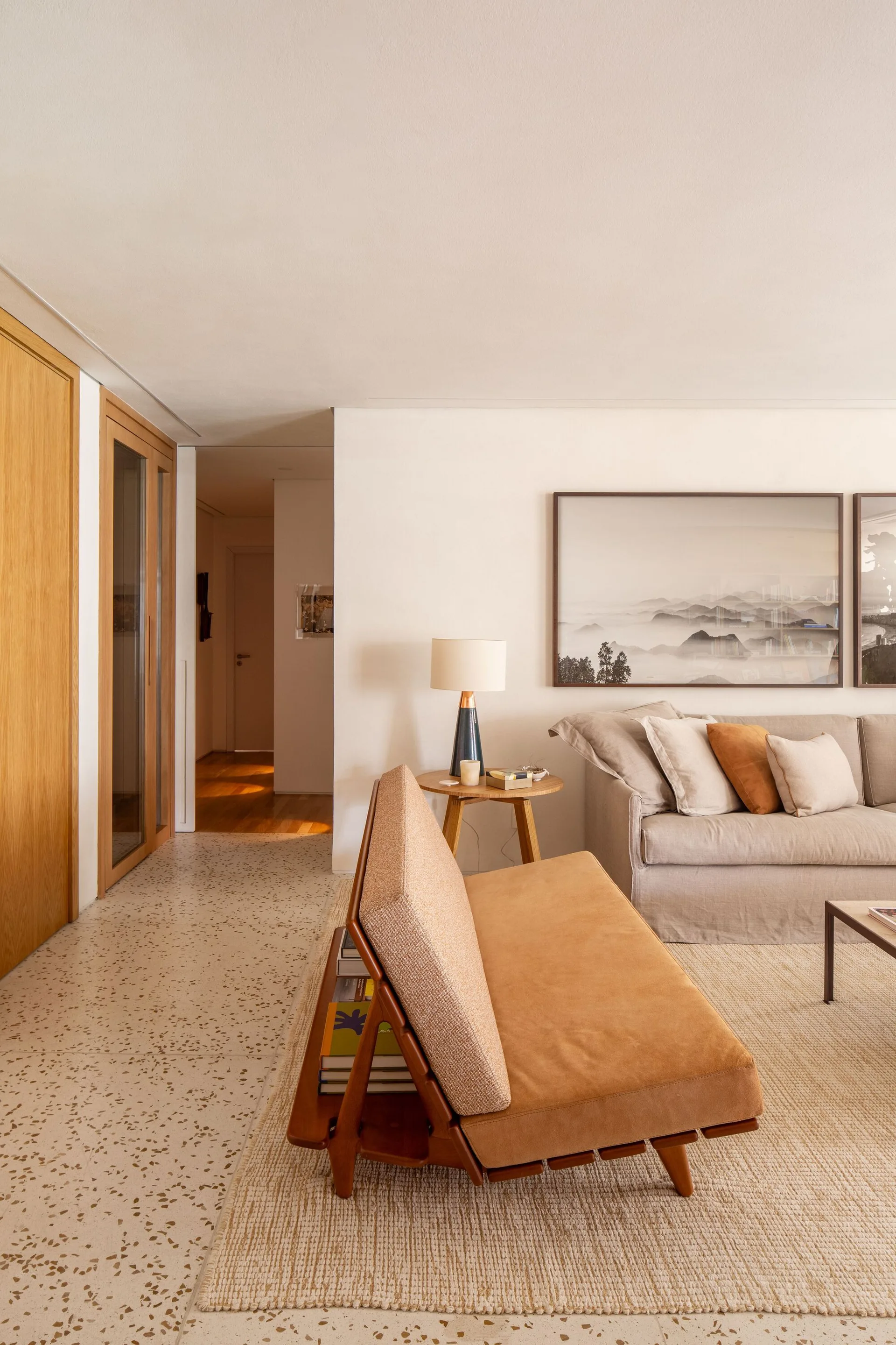 A modern living room with a tan suede daybed, a beige linen sofa, a wooden side table with a lamp, and speckled terrazzo flooring.
