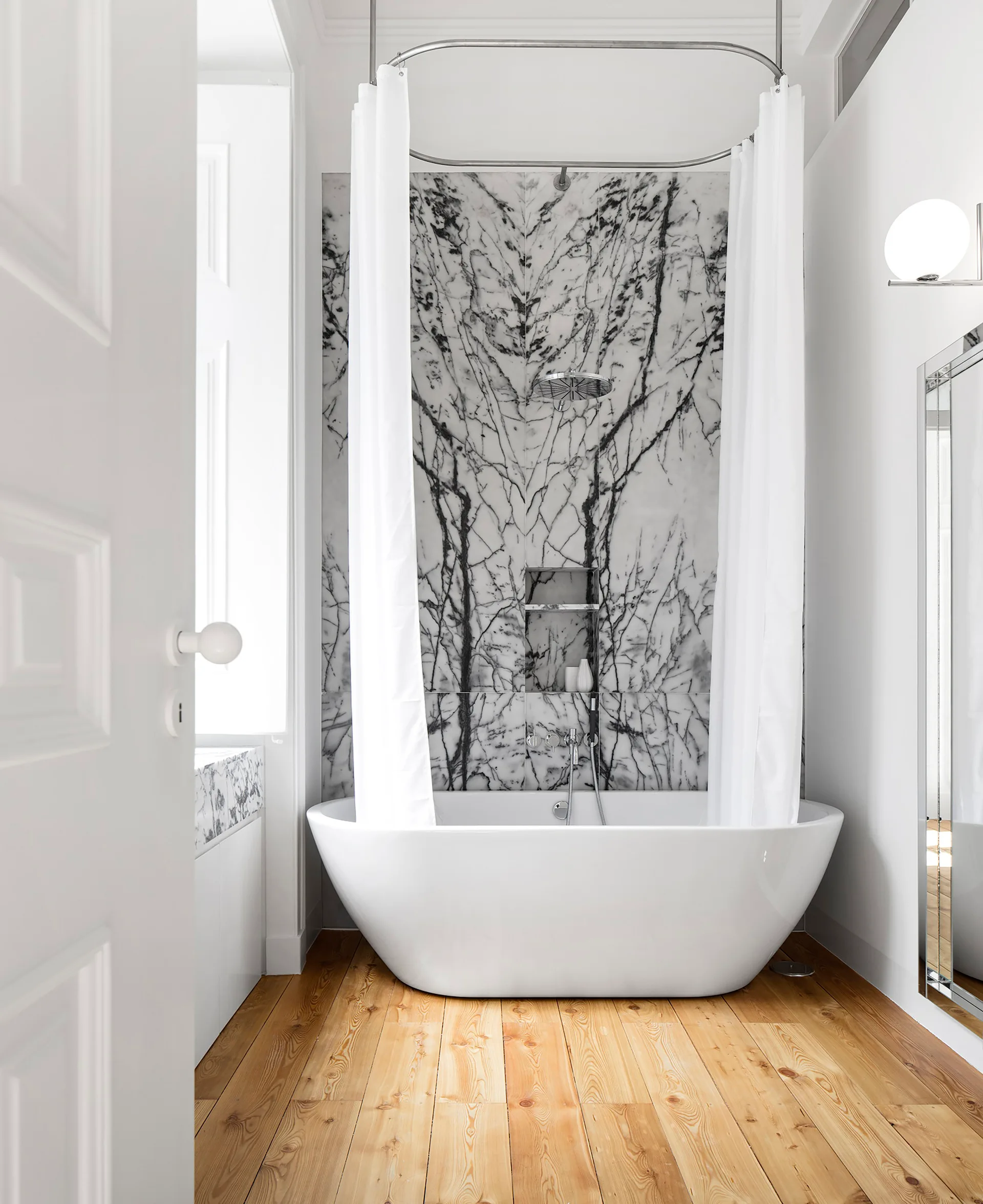 A white freestanding bathtub sits on wood plank flooring in front of a dramatic black and white veined marble accent wall with a suspended shower curtain.