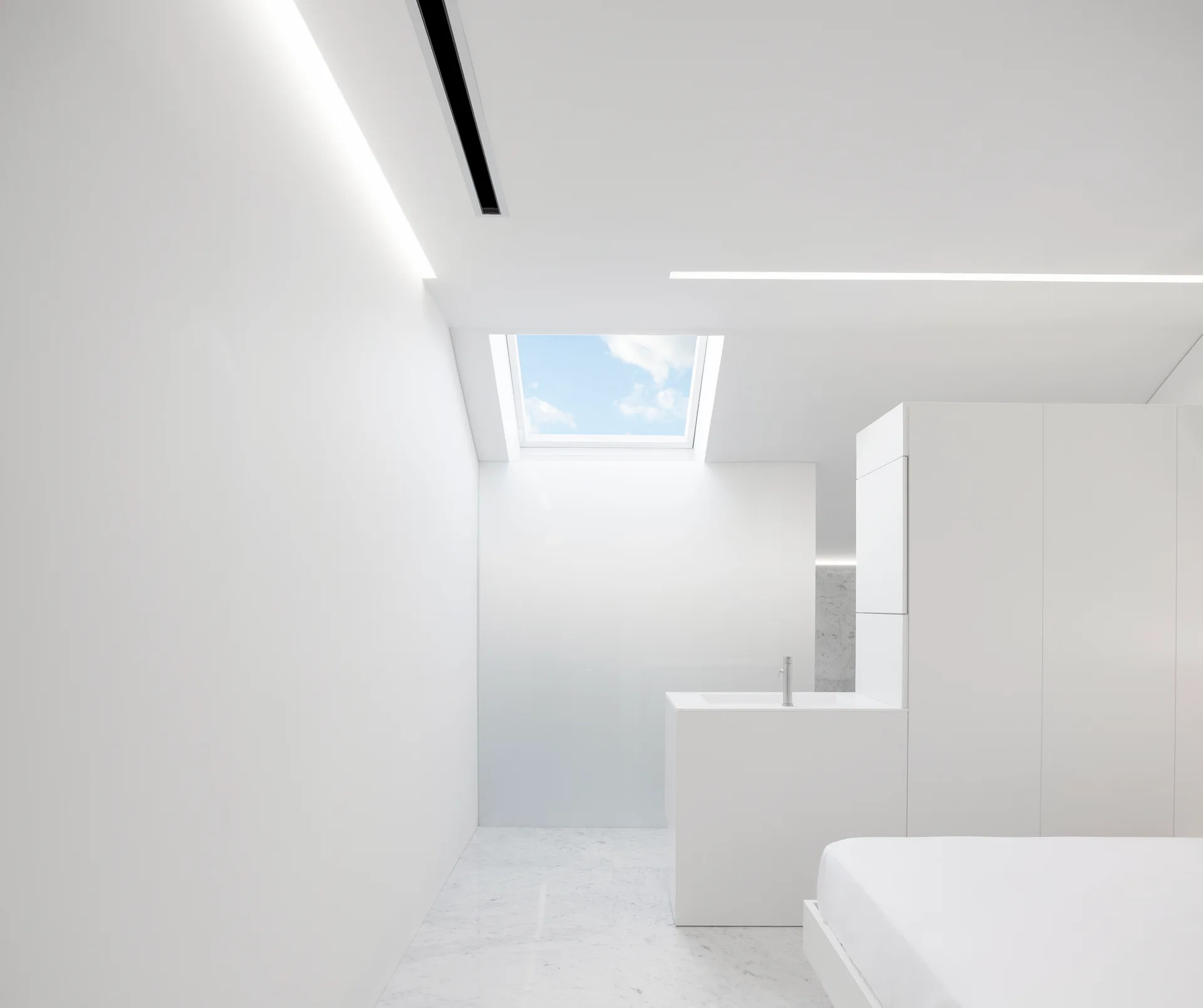 A minimalist white interior space featuring a monolithic vanity, a skylight showing blue sky, linear ceiling lights, and marble flooring.