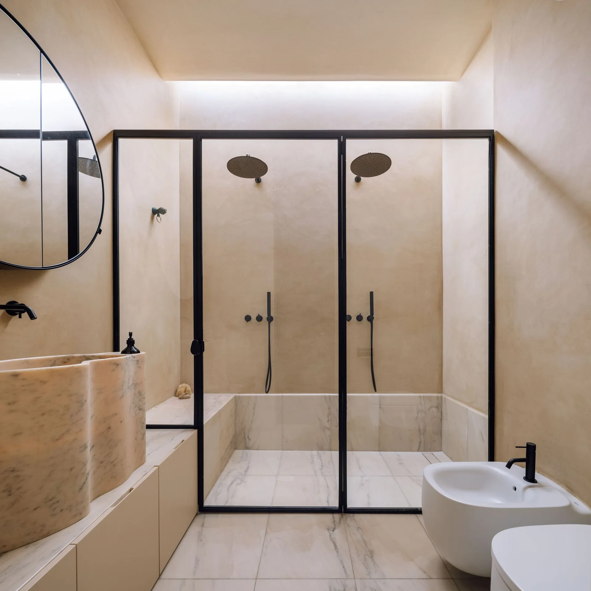 A contemporary beige bathroom features a black-framed walk-in double shower, a sculptural curved marble sink, and a matching bidet.