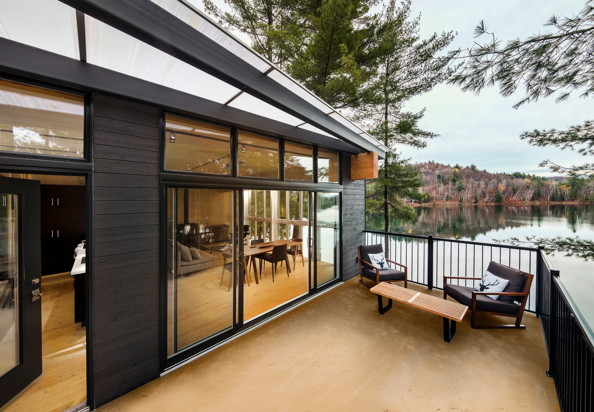A modern lakeside terrace with dark charcoal horizontal siding, two lounge chairs, a slatted wood coffee table, and large sliding glass doors.