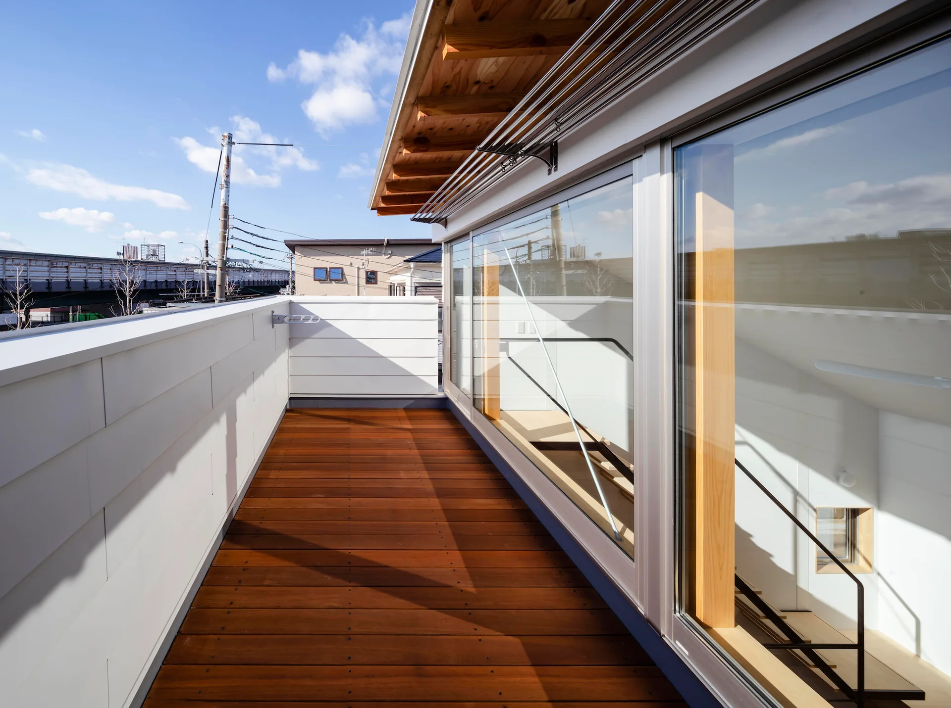 An outdoor wooden balcony deck features a solid white panel railing and a large sliding glass wall showing an interior staircase.