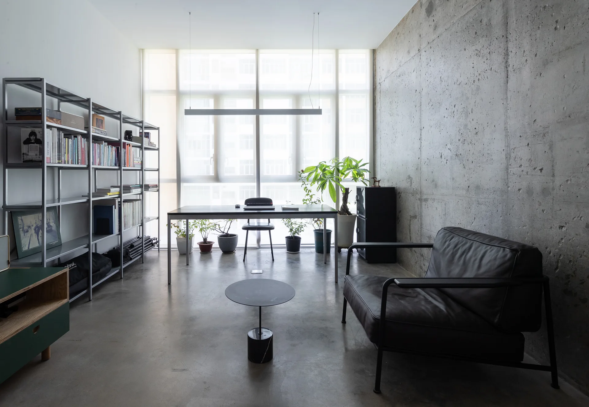 A minimalist home office featuring a textured concrete wall, a metal bookshelf, and a desk positioned in front of a large window.