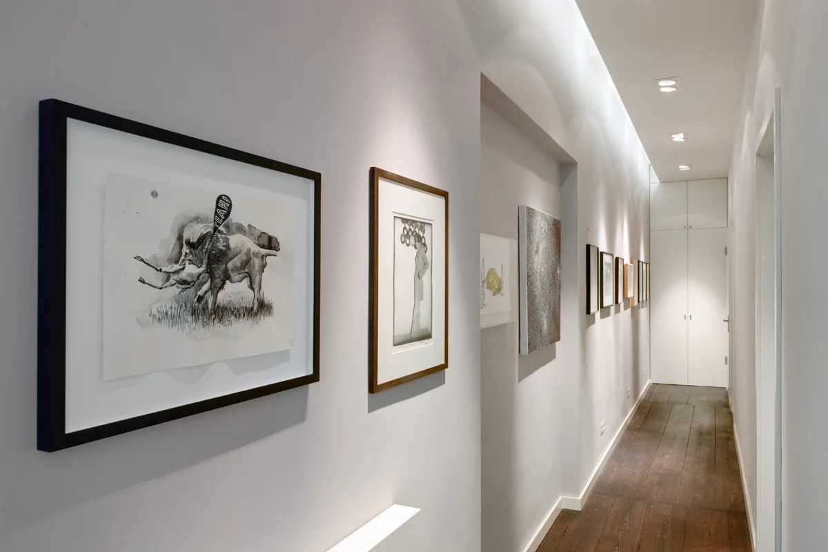 A long white residential hallway features framed artwork on the walls, dark wood flooring, and recessed ceiling spotlights.