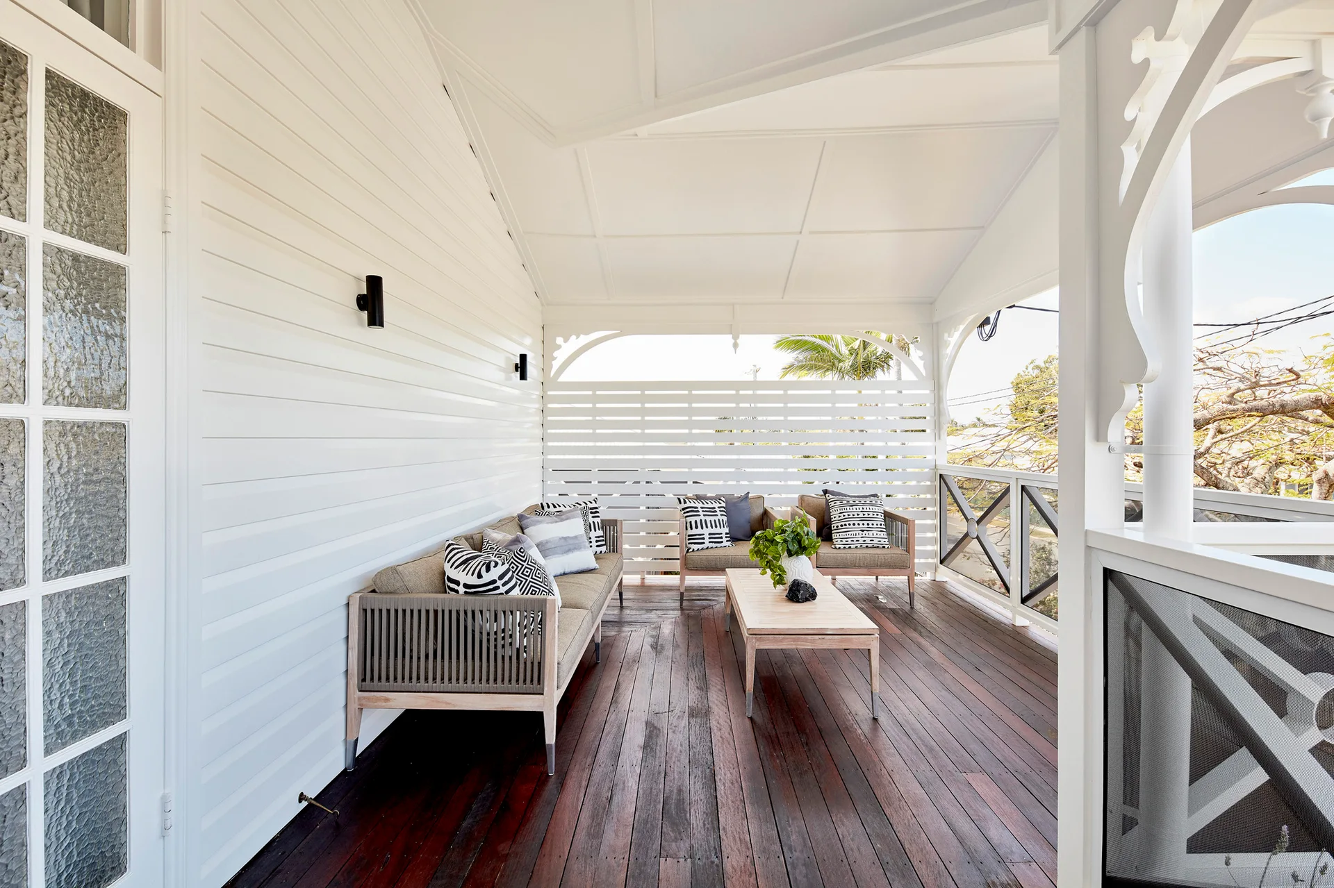 A covered wooden veranda features a grey cushioned sofa, matching armchairs, and a low coffee table set against white horizontal wall paneling.