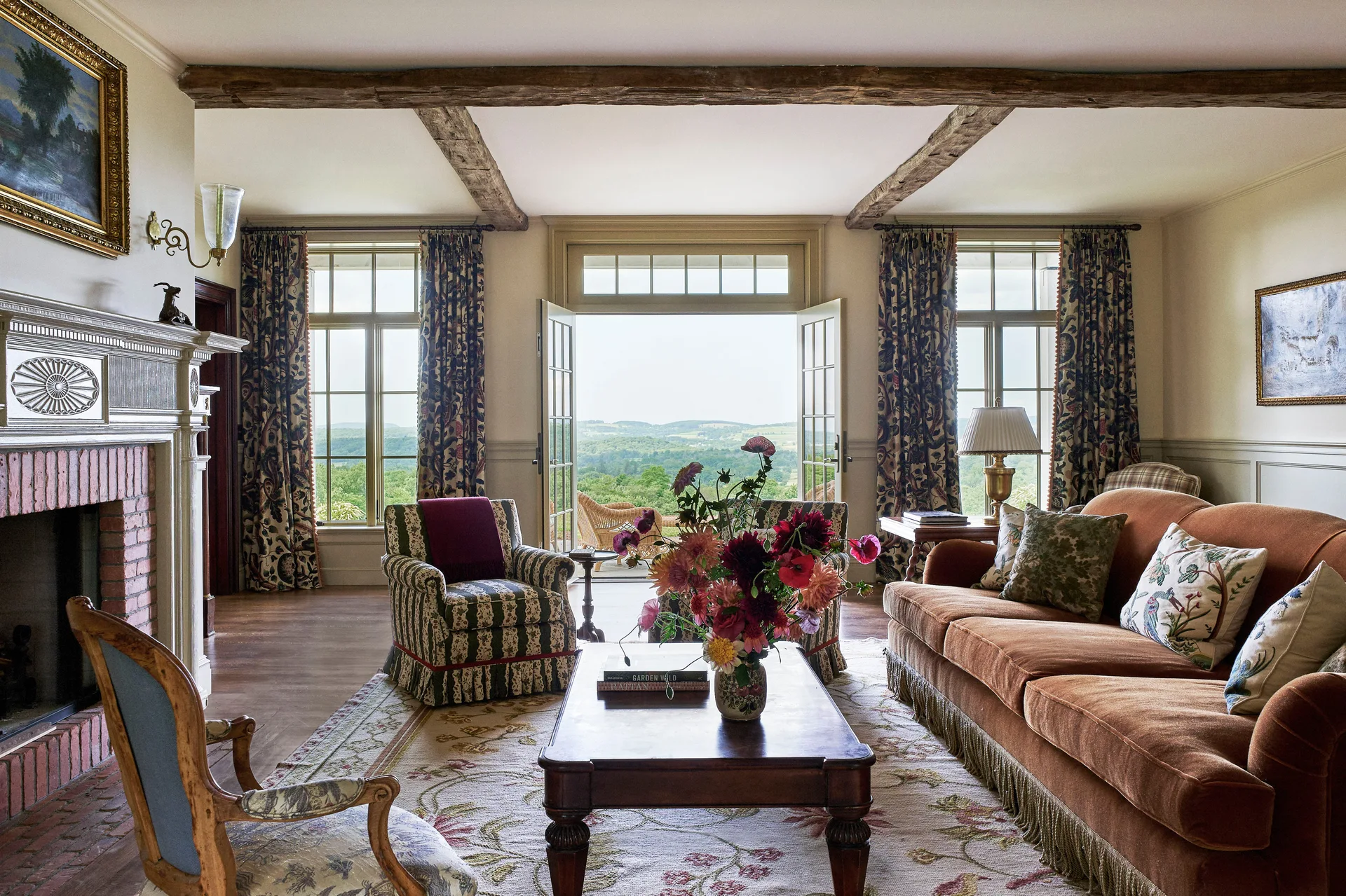 A traditional country living room with a terracotta velvet sofa, exposed ceiling beams, a brick fireplace, and open French doors showcasing a green landscape.