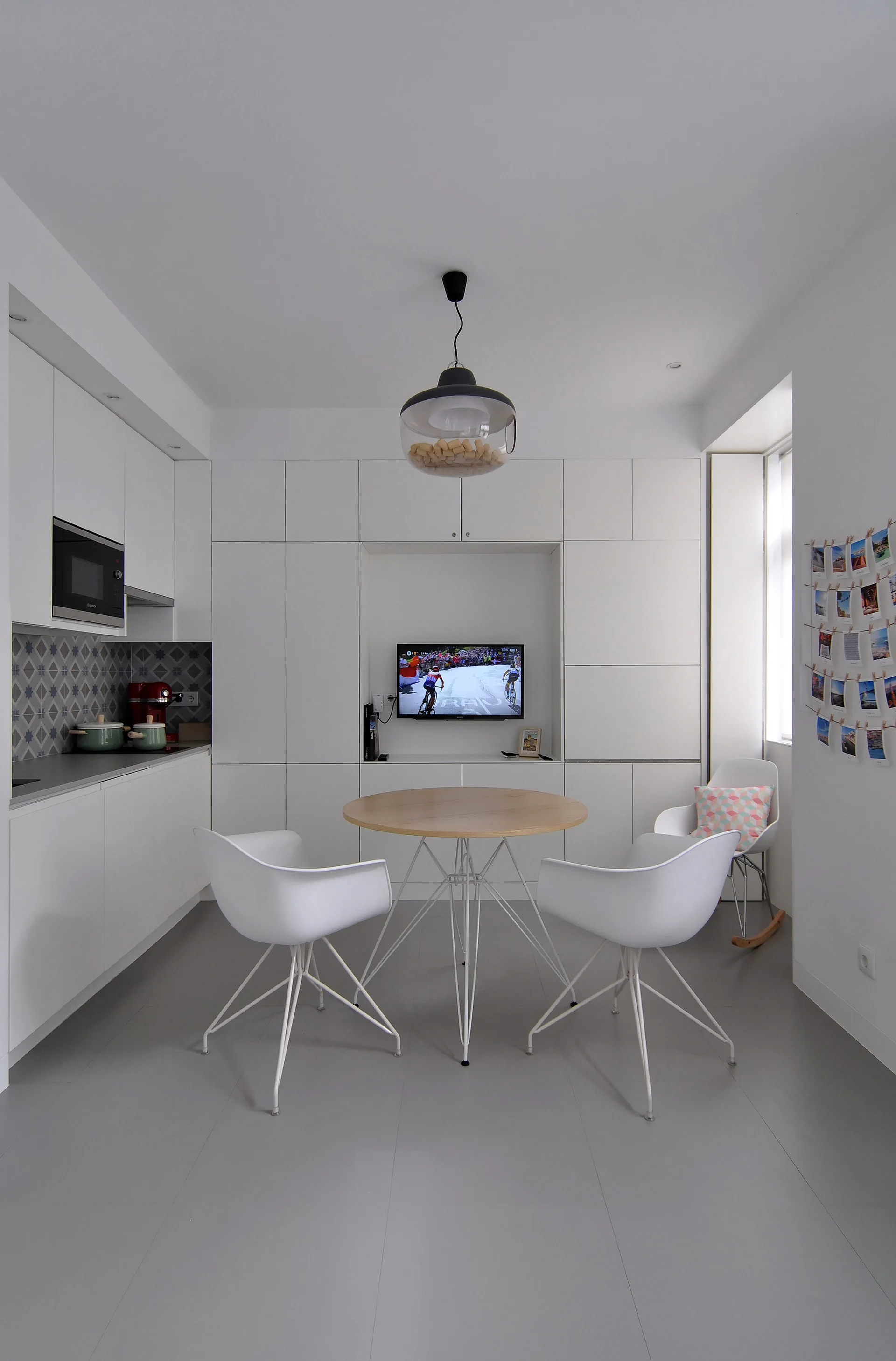 A modern white kitchen features seamless built-in cabinets, a recessed television niche, and a round wooden dining table surrounded by white chairs.
